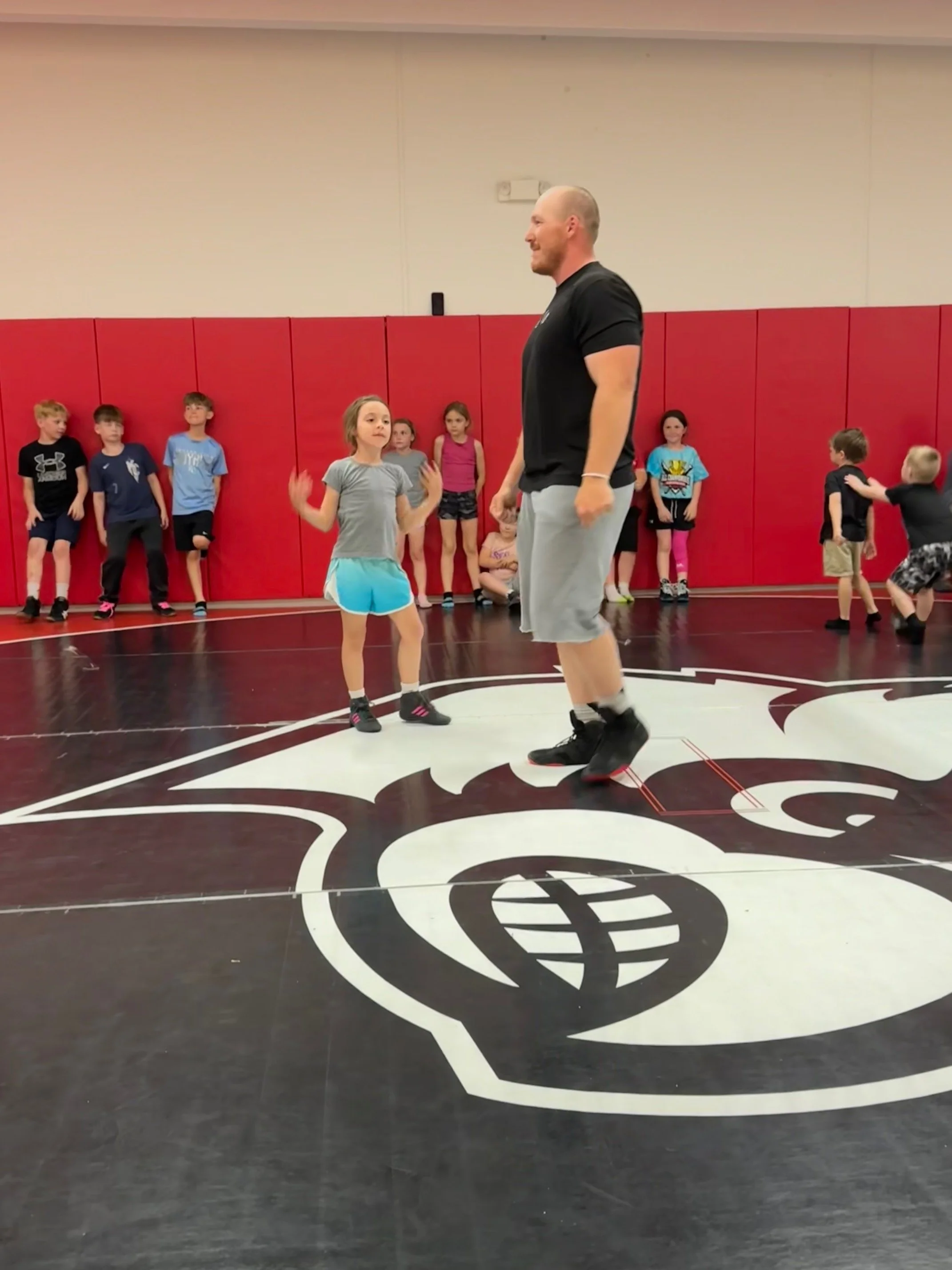 A man teaching children basketball drills on a gymnasium floor with a black and white NBA logo.