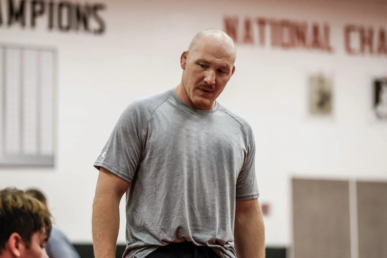 A bald man wearing a gray t-shirt standing in a gym or sports facility, with a focused expression.
