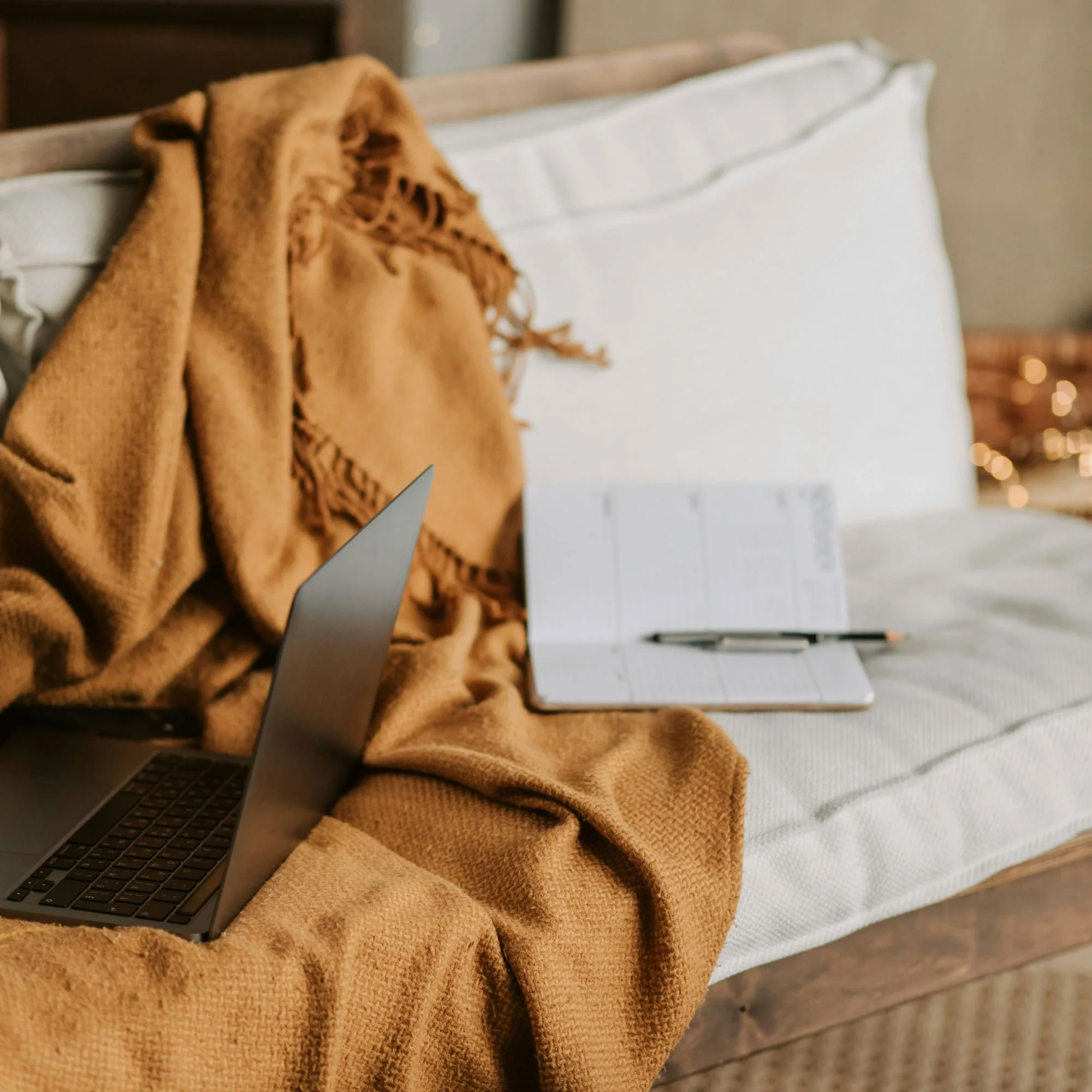 Close-up of a cozy scene with a laptop resting on a soft, caramel-colored blanket, with a white pillow and a blurred background of fairy lights.