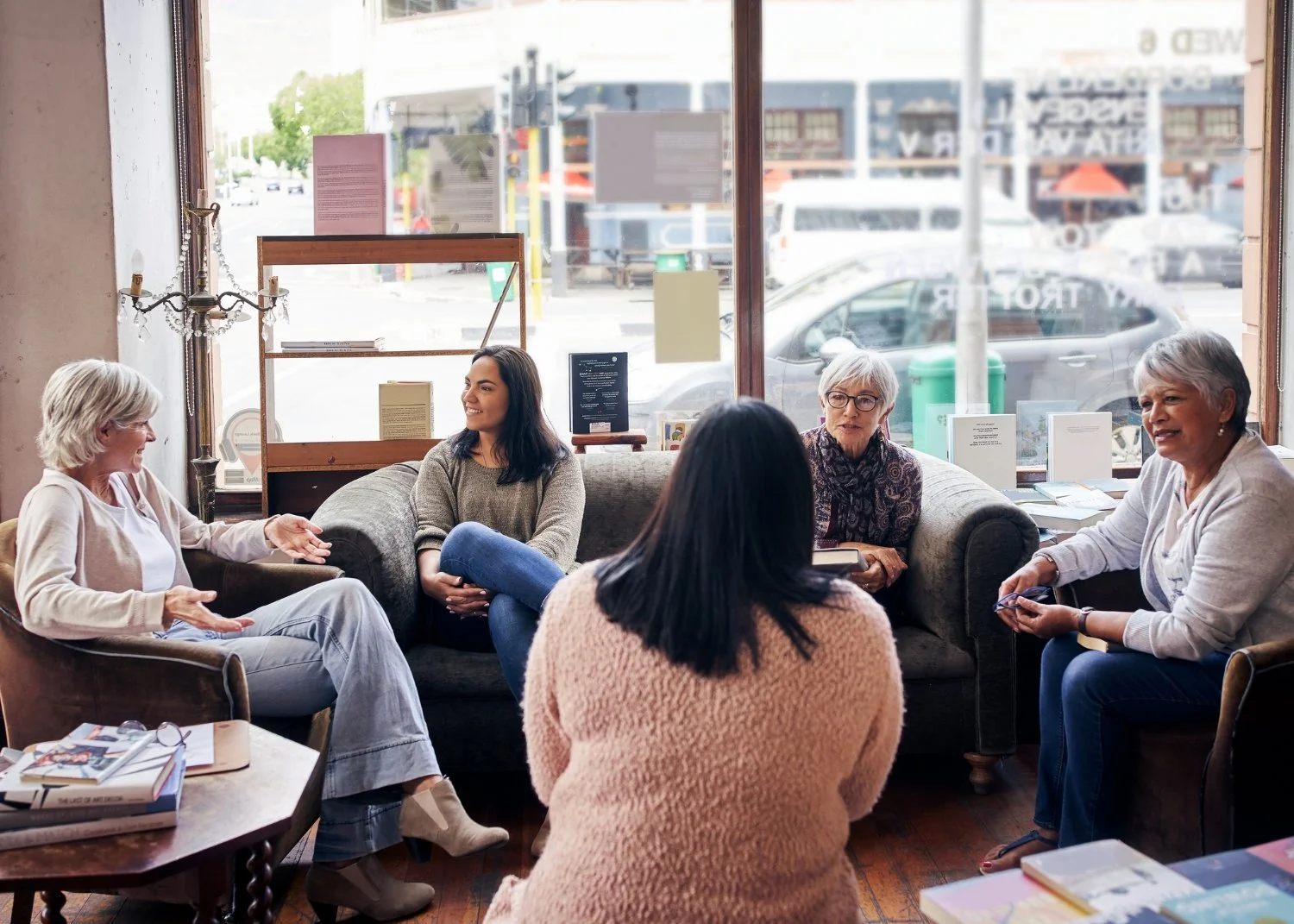 Five women having a conversation in a cozy cafe or bookstore, sitting on sofas and chairs, with books and papers on a table in the foreground and large windows showing a street scene outside.