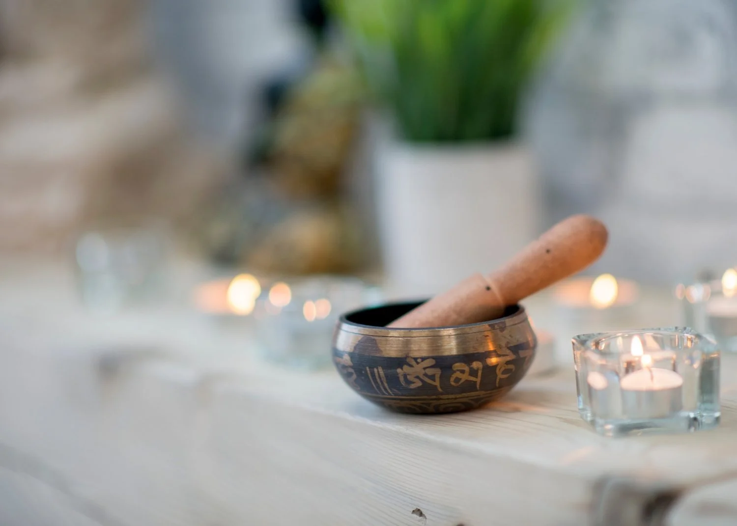 A Tibetan singing bowl with a wooden striker inside, surrounded by lit candles, with a blurred background of potted plants.