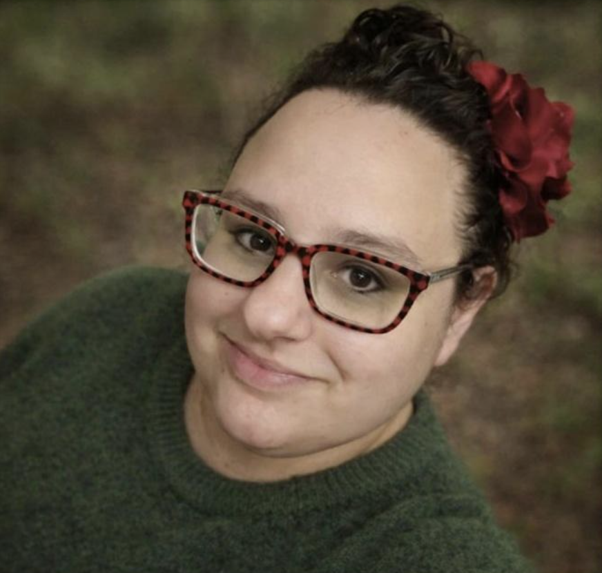 A woman with glasses, dark curly hair with a red flower accessory, wearing a green sweater, smiling outdoors.