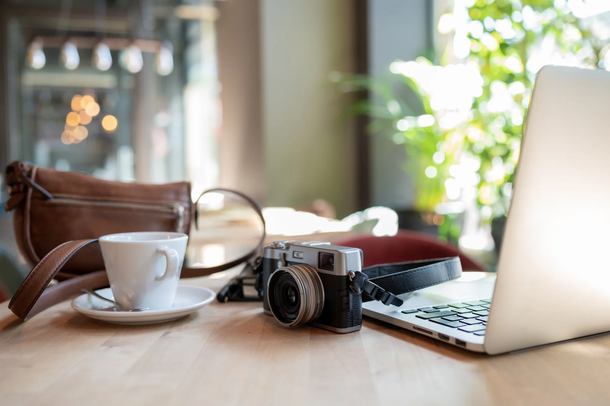 A workspace with a vintage camera, a white coffee cup on a saucer, a brown leather bag, a laptop, and a camera strap on a wooden table. In the background, there is greenery outside the window and hanging lights inside the room.