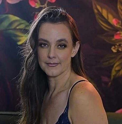 A woman with long dark hair wearing a sleeveless top, looking at the camera, in front of a decorative background with leaves and flowers.