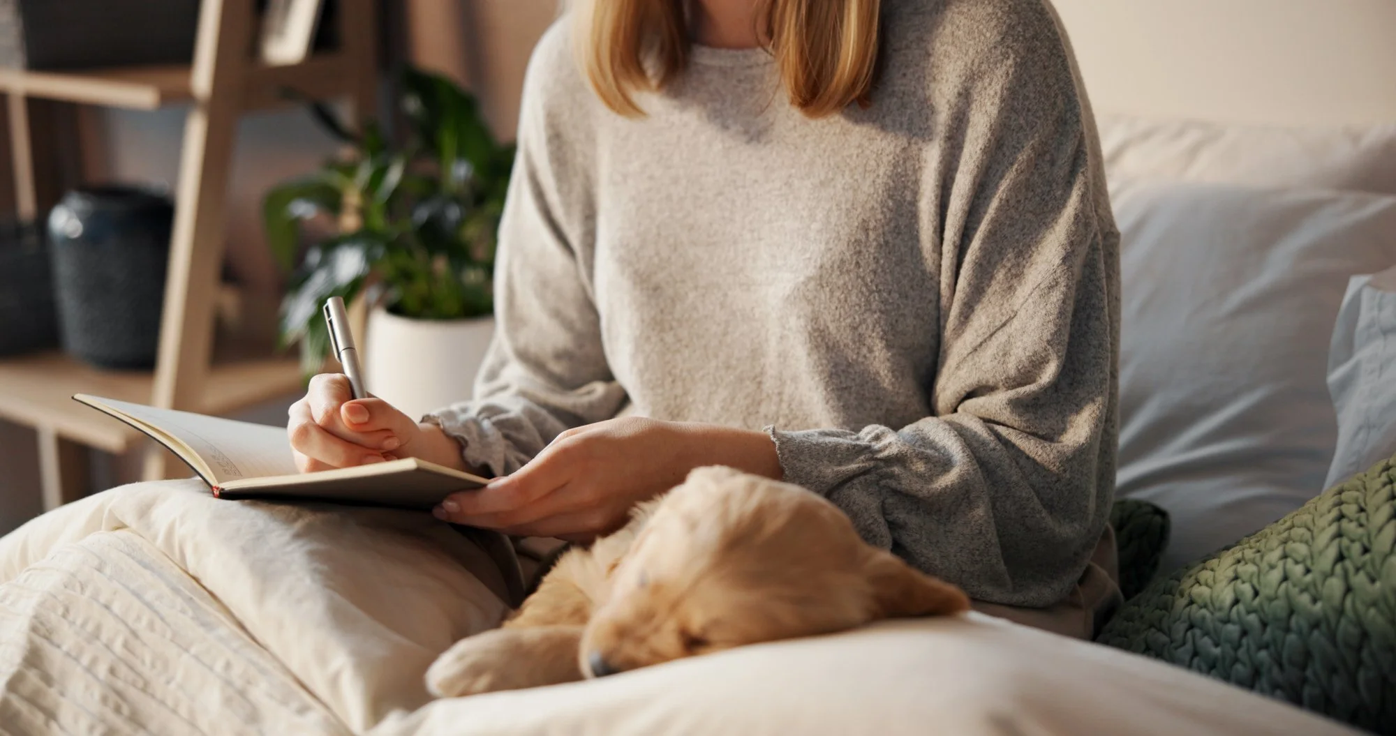 A person with red hair sitting on a bed with a beige quilt, writing in a notebook with a pen, a yellow dog resting nearby, a bookshelf and a potted plant in the background.