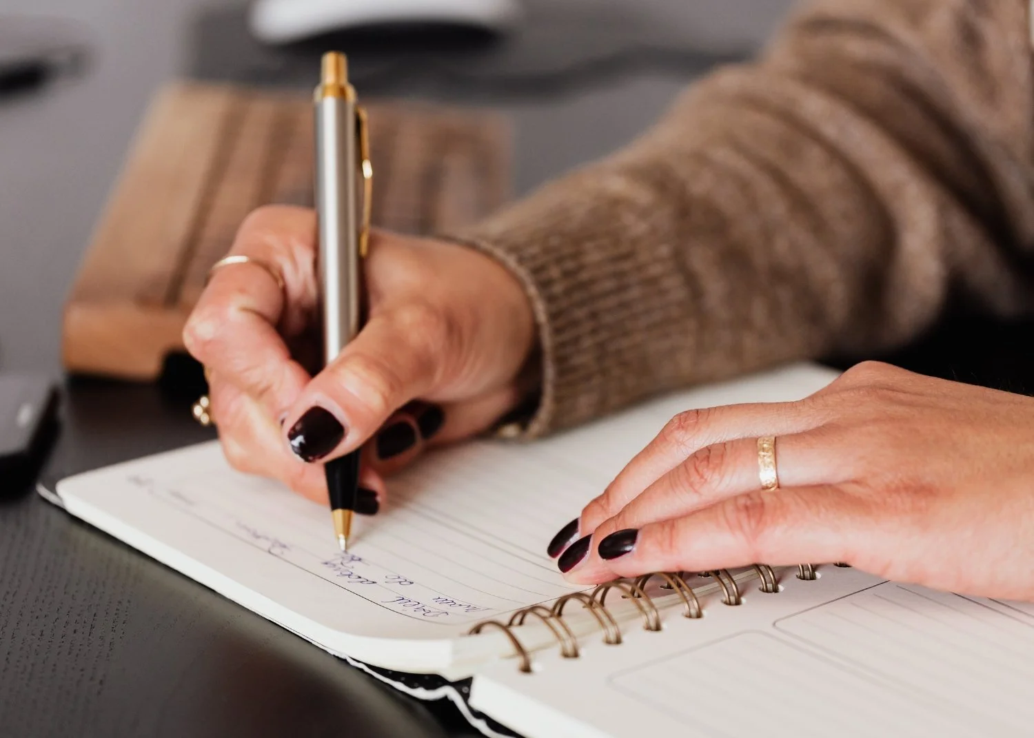 A person writing in a notebook with a pen, with their hand resting on the page. The person has black nail polish and is wearing a gold ring. The background shows a laptop and a wooden object.