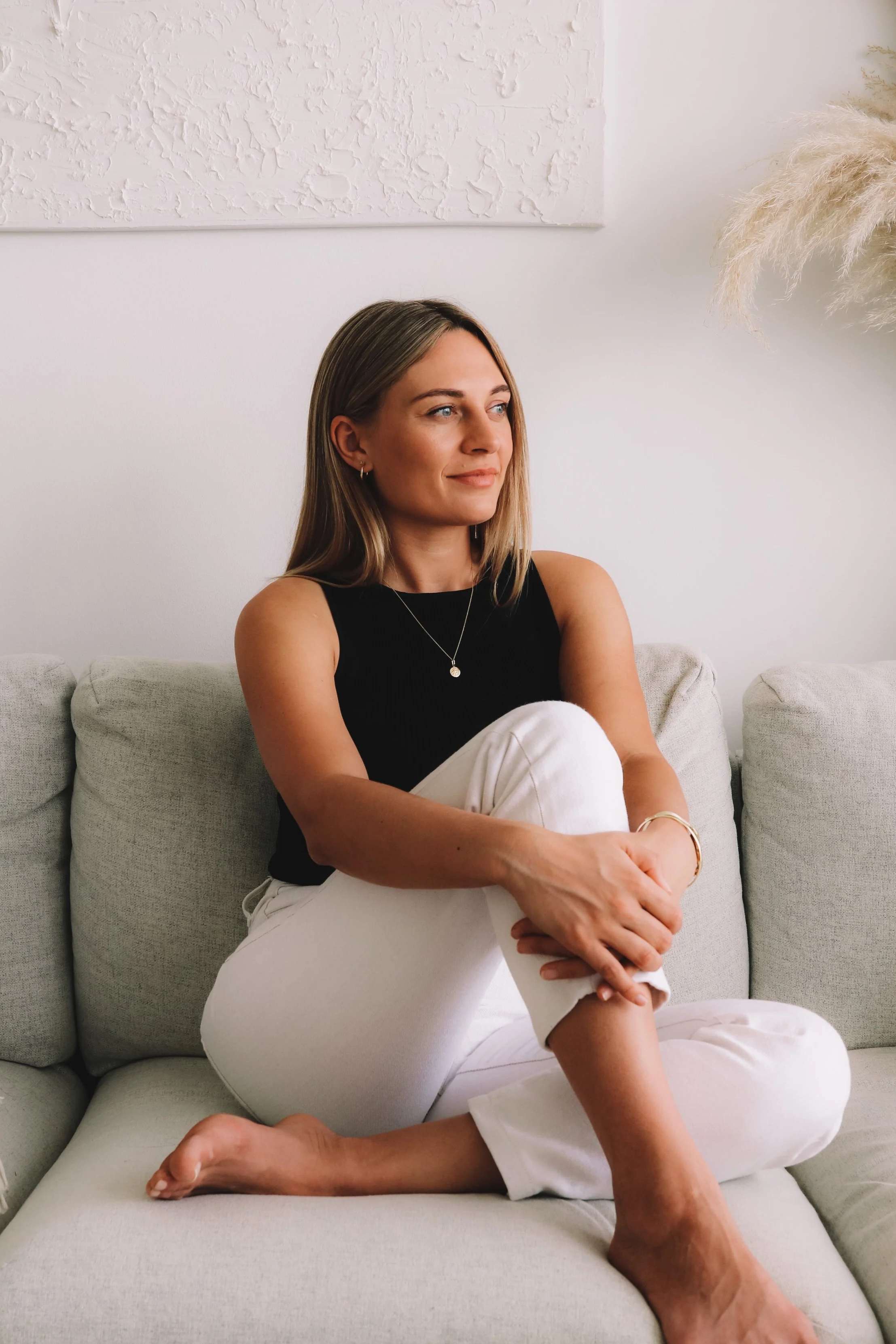 A woman with blonde hair, wearing a black sleeveless top, sitting cross-legged on a light-colored sofa in a minimalistic room with textured white walls and a decorative potted plant.