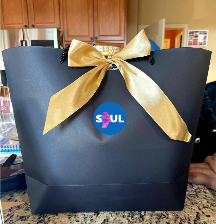 A black gift bag with a large gold ribbon bow on the front. The bag has a round blue sticker with the word 'SOUL' and a purple face illustration.