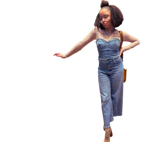 A woman with curly hair wearing glasses, a white long-sleeve top, a denim bustier, high-waisted jeans, and heels, posing with her hand on her hip against a plain background.