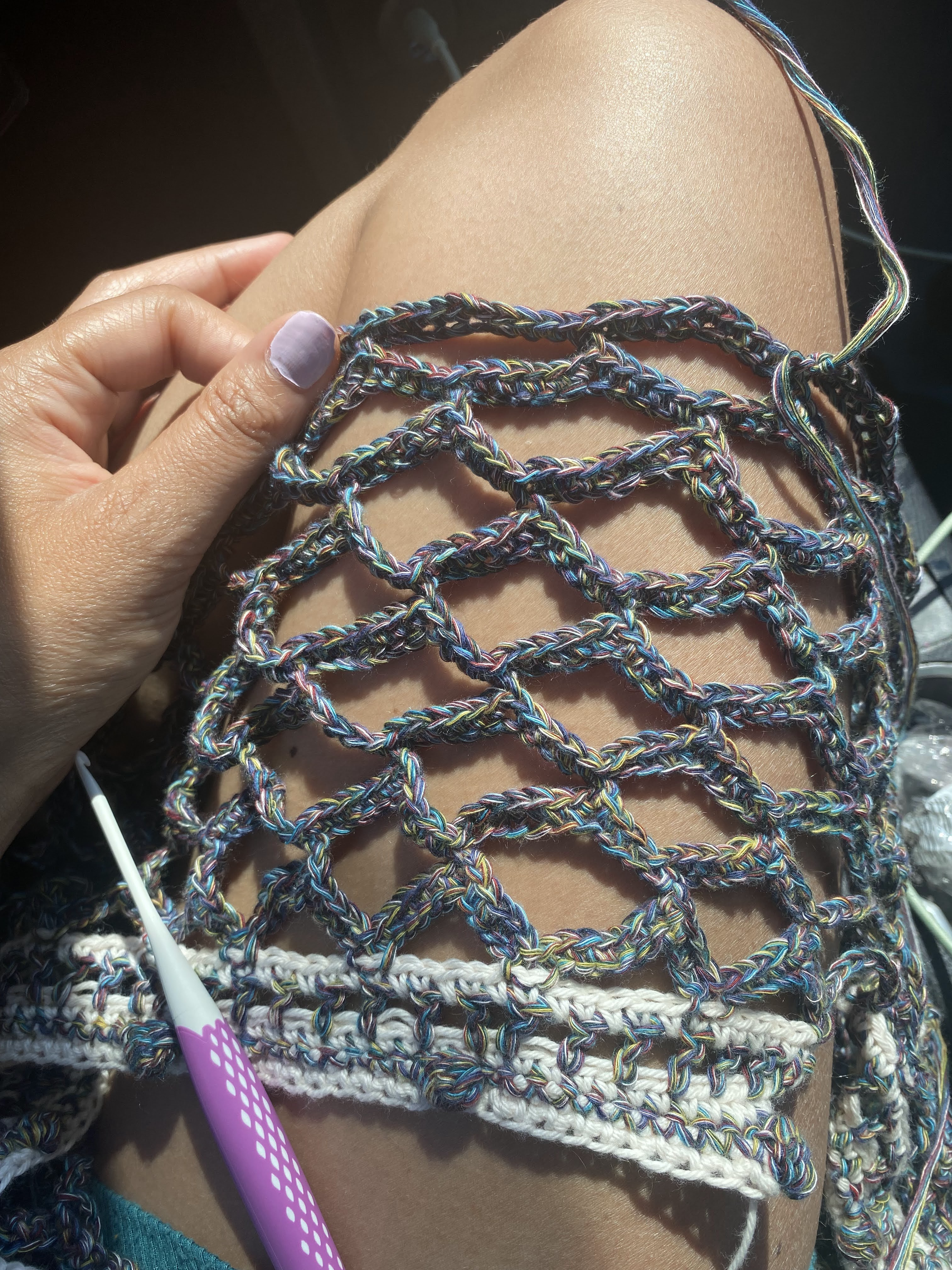 Close-up of a person's knee with a detailed view of colorful, handcrafted crochet skirt in progress and a purple ergonomic crochet hook resting on top of the skirt.