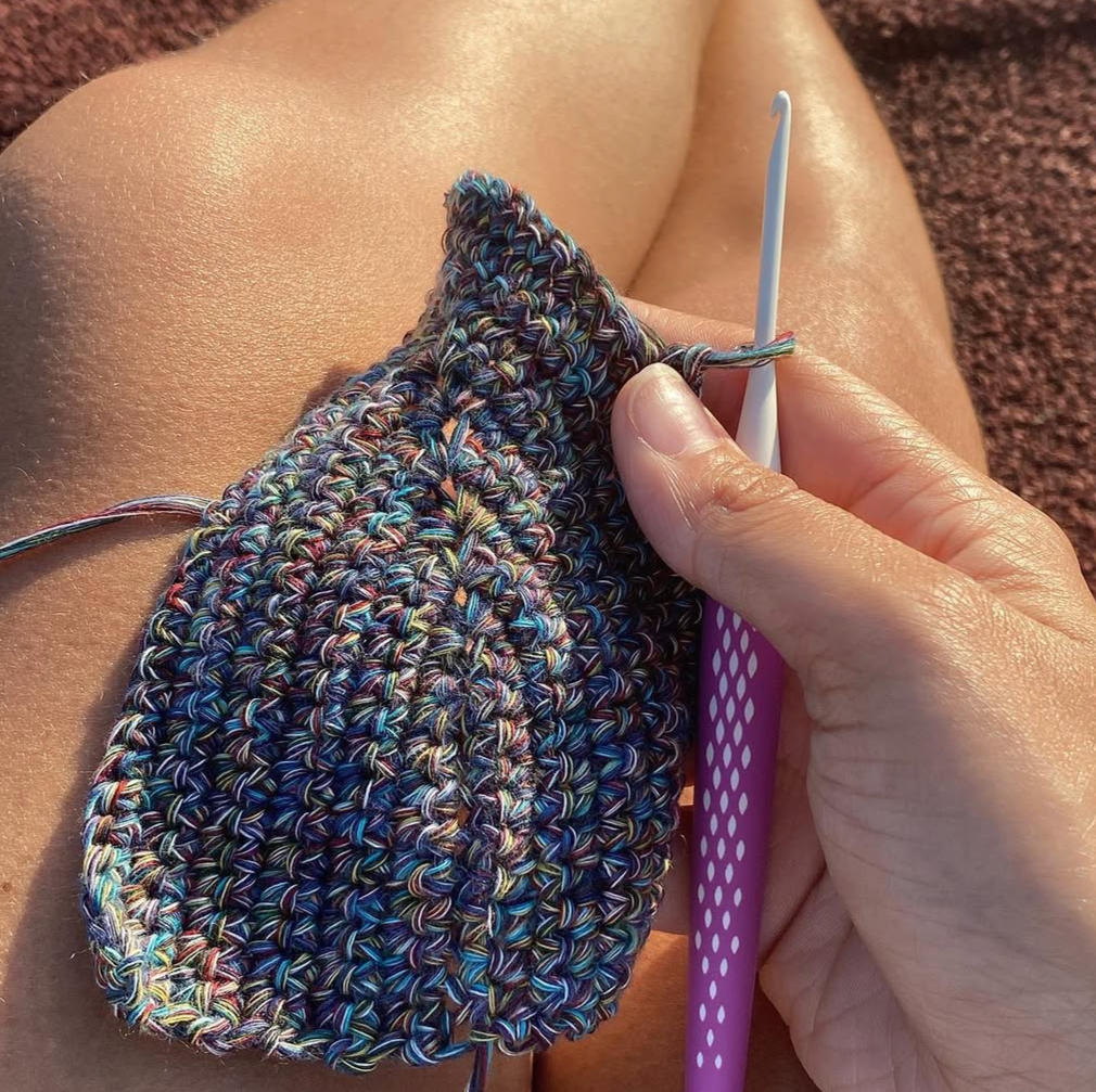 Close-up of a person crocehting a colorful, multi-hued piece with a purple crochet hook, sitting outdoors on a reddish-brown surface.