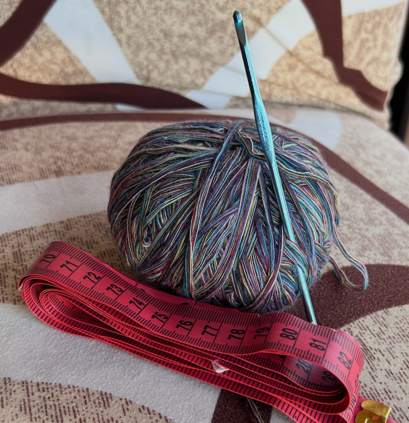 A multicolored ball of yarn with a crochet hook inserted into it, placed on a striped fabric surface alongside a pink measuring tape.