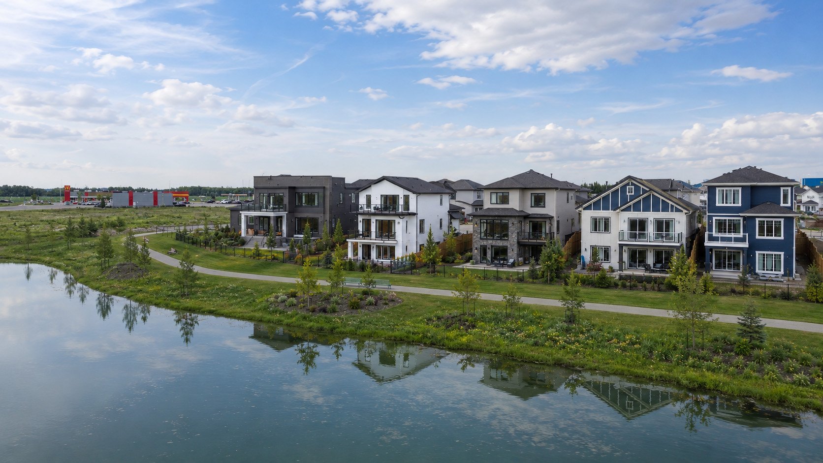 A row of modern houses with balconies and large windows, situated along a curved path beside a calm lake, with greenery and trees in the foreground and a partly cloudy sky overhead.