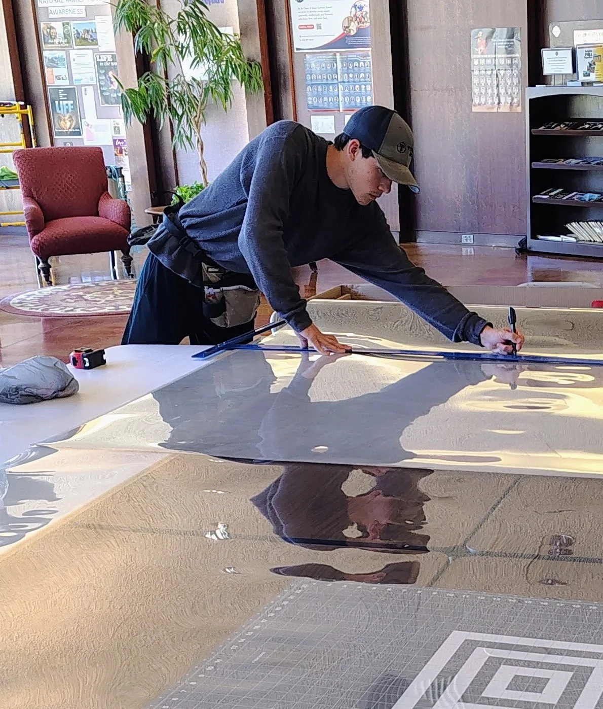 A person wearing a cap and dark clothing is measuring a large, reflective surface, possibly a metal sheet or similar material, on a table in an indoor setting with wooden floors and walls decorated with posters and a bookshelf.