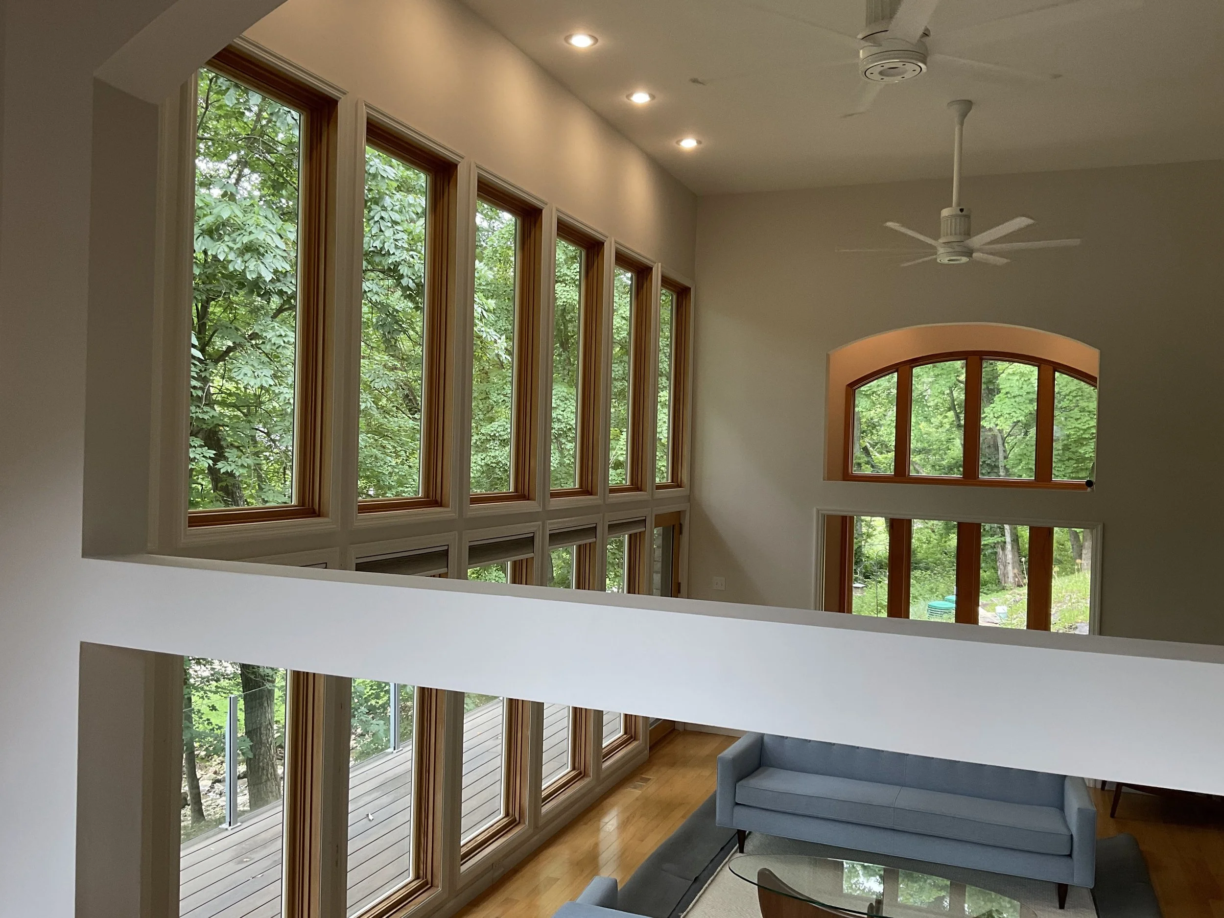A living room with large windows overlooking green trees, hardwood floors, a white ceiling with recessed lights, and a white ceiling fan. There's a gray couch and a glass coffee table.