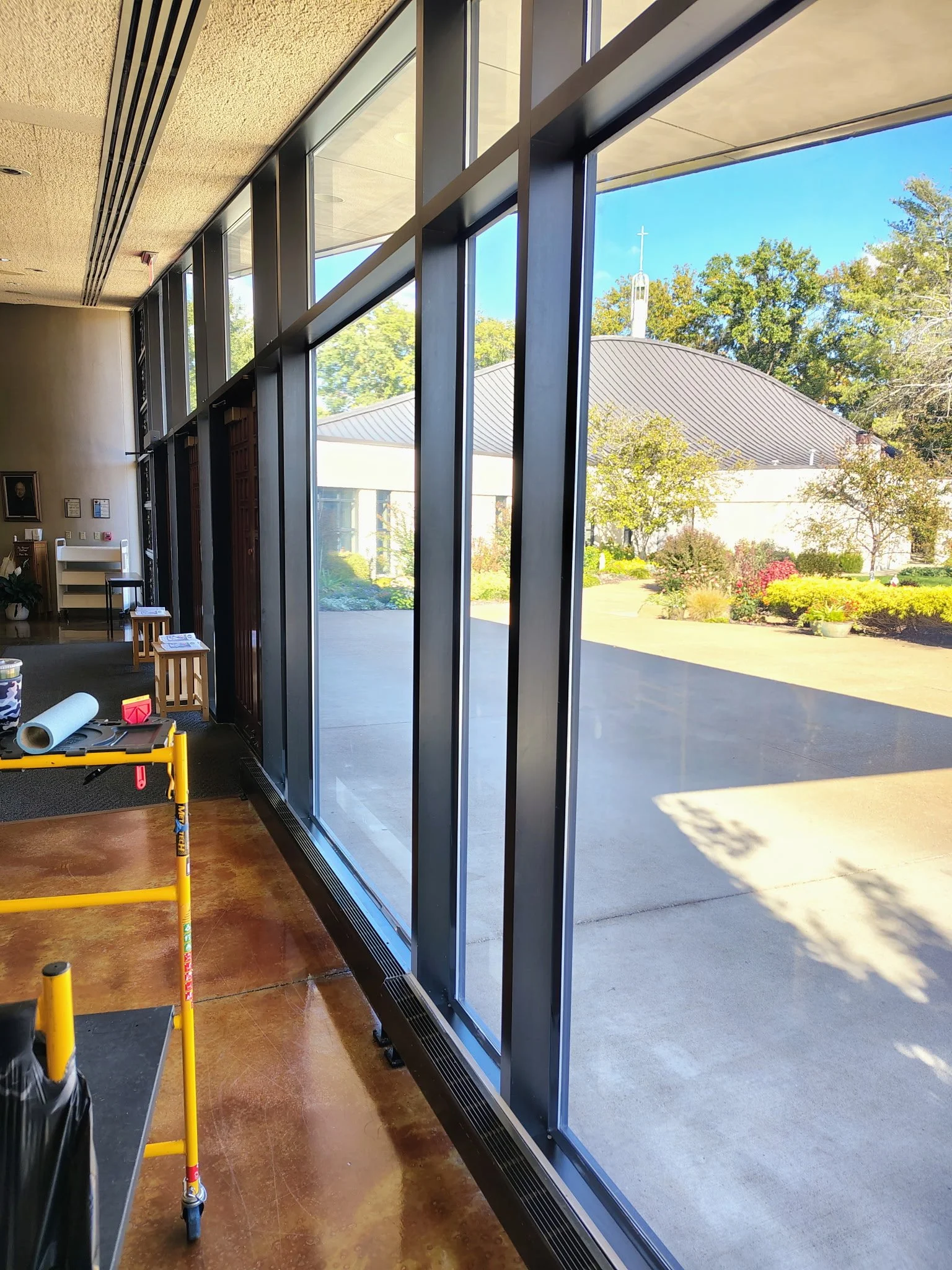 Interior view of a large modern building with tall floor-to-ceiling windows showing a landscaped garden outside, including trees, bushes, and a building with a steeple. Inside, there are some small stools, a yellow tool cart, and construction or main