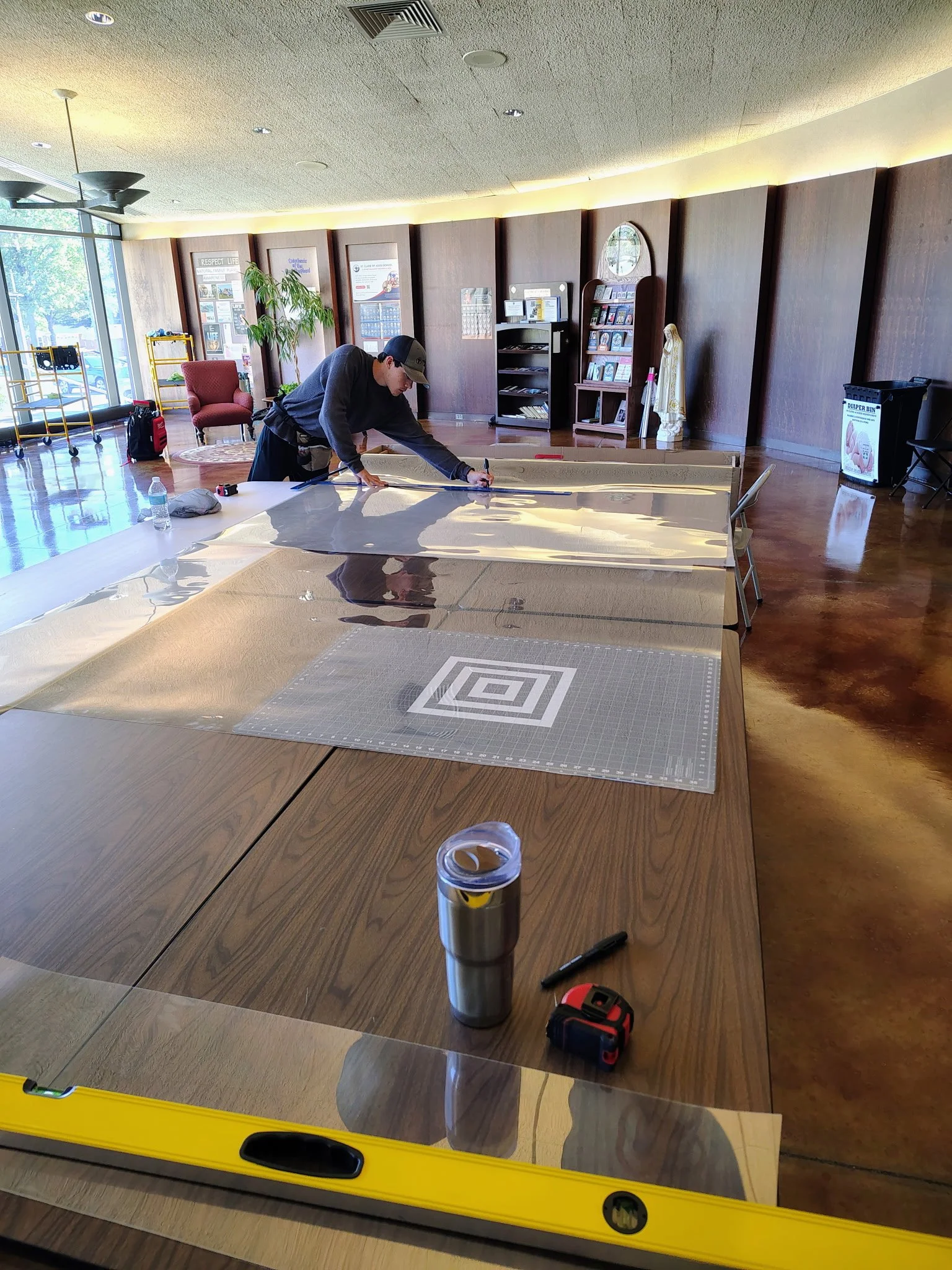 A man in a black cap and dark clothing working on a large glass panel indoors. The room has wooden floors, a large window, and various items like a chair, a potted plant, shelves with pamphlets, and a statue. Tools and a water bottle are on the table
