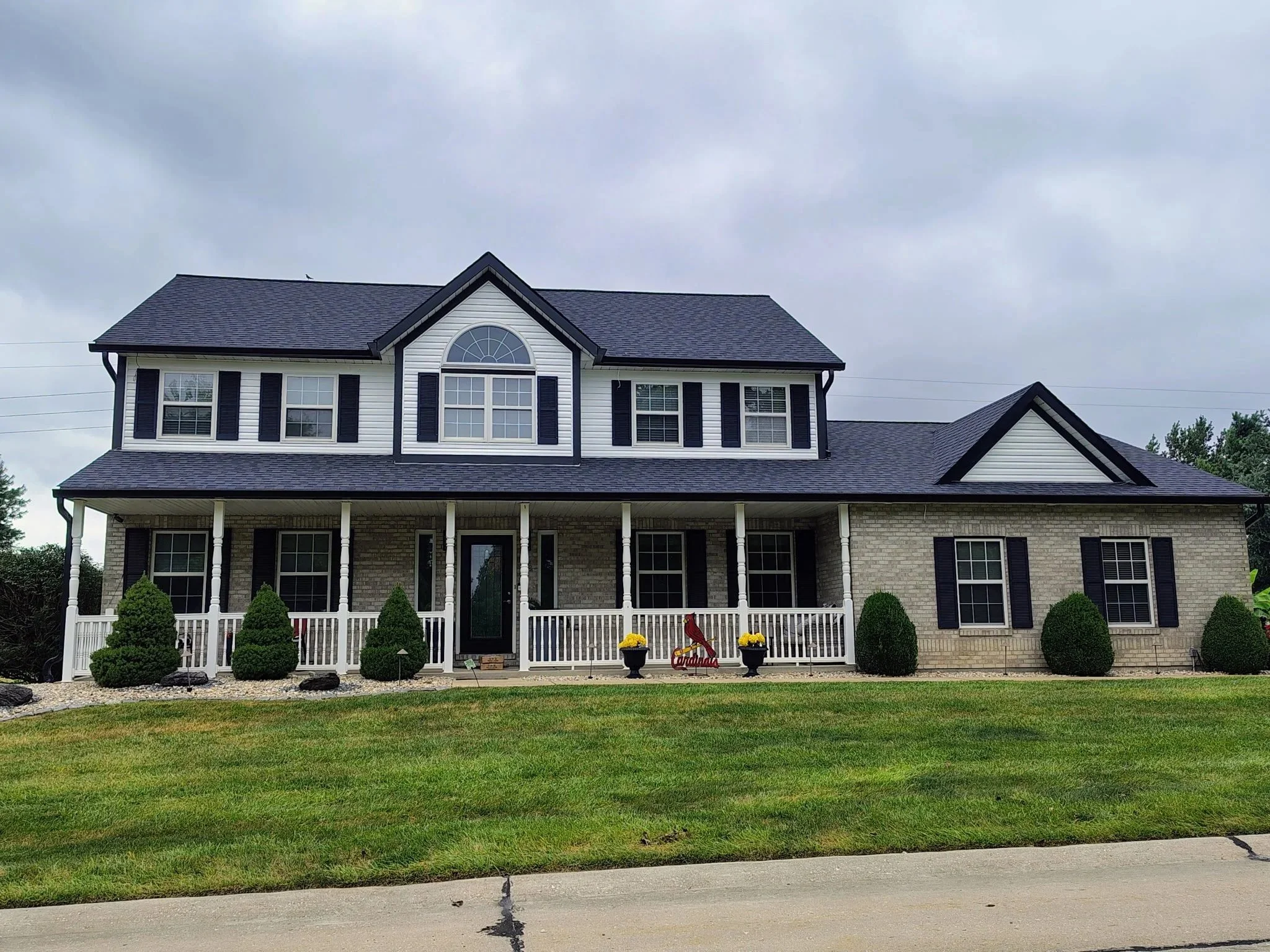 A two-story house with white siding, black shutters, and a dark roof. It has a front porch with white columns and a brick foundation. There are small bushes and decorative flowers in planters outside, and a lawn with a sidewalk in front. The sky is o