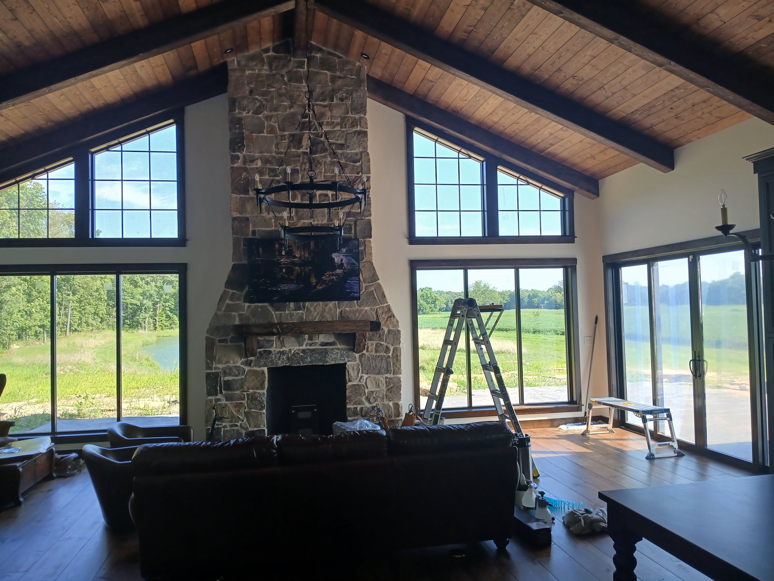 Living room with large windows, a stone fireplace, a black chandelier, a TV above the fireplace, a ladder, and various furniture and construction items inside.