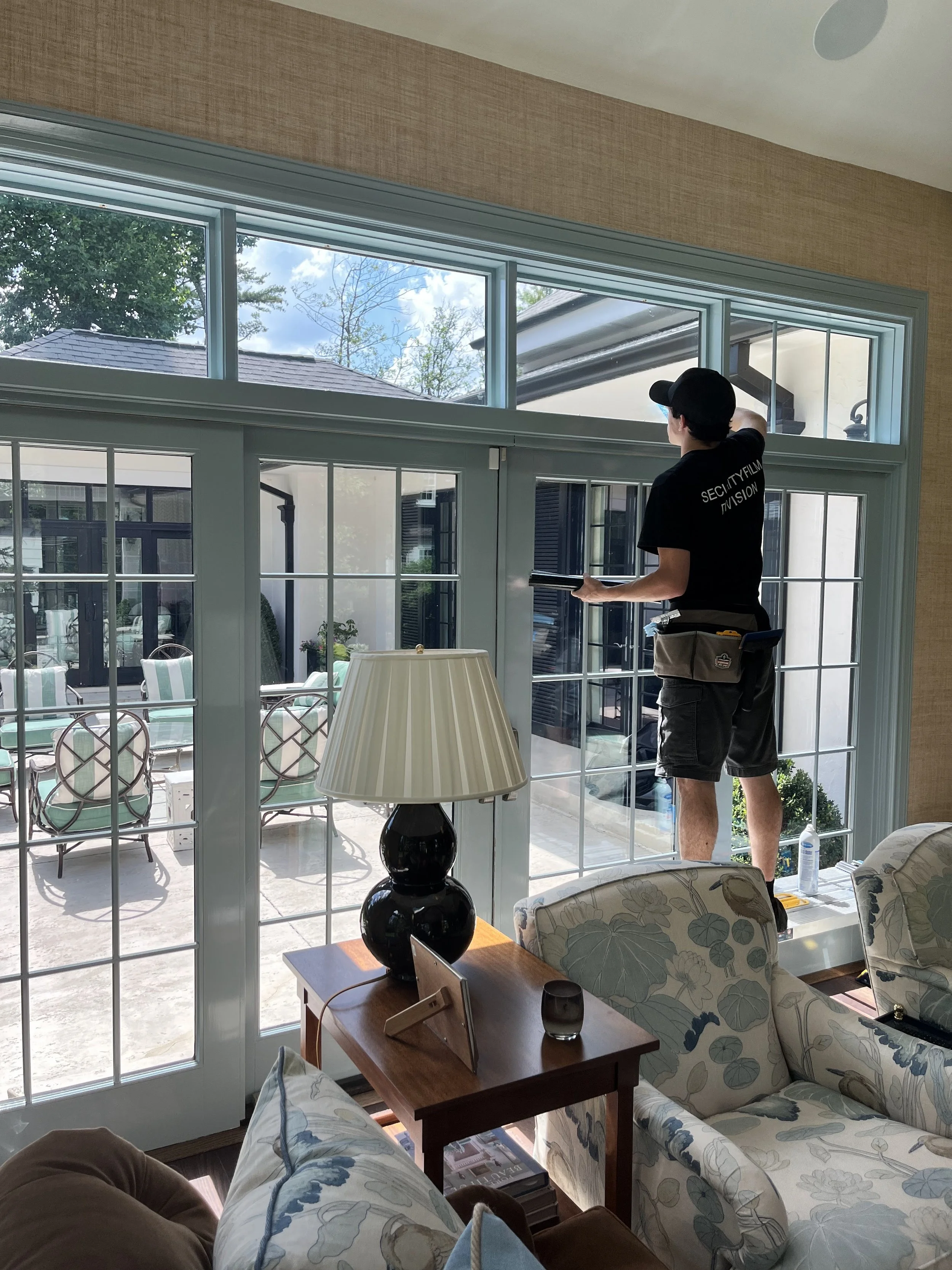 A man wearing a black T-shirt and cap is cleaning large windows in a living room. The room has a sofa with floral pattern upholstery, a wooden side table with a black lamp and some items, and a view outside showing patio furniture and a house with trees.