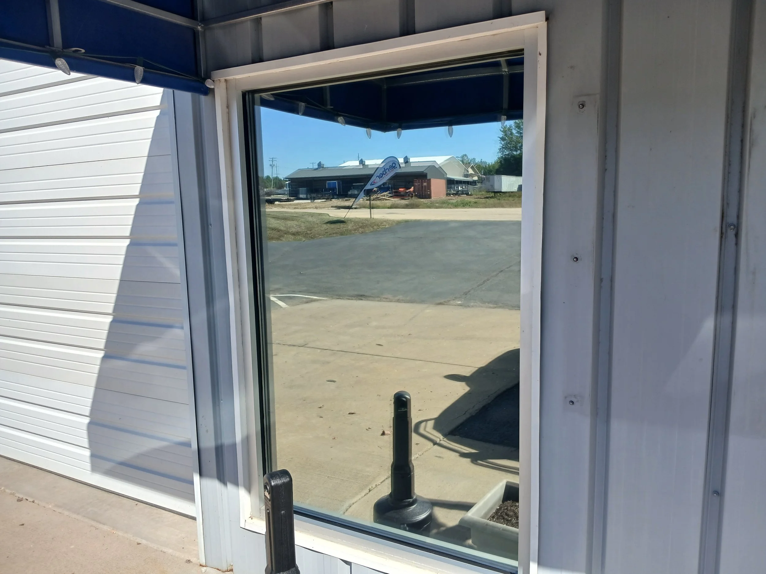 View through a missing or open glass door to a parking lot with a small building in the background and a blue flag sign. The building appears under construction, with construction materials around. The image is taken during daytime on a sunny day.