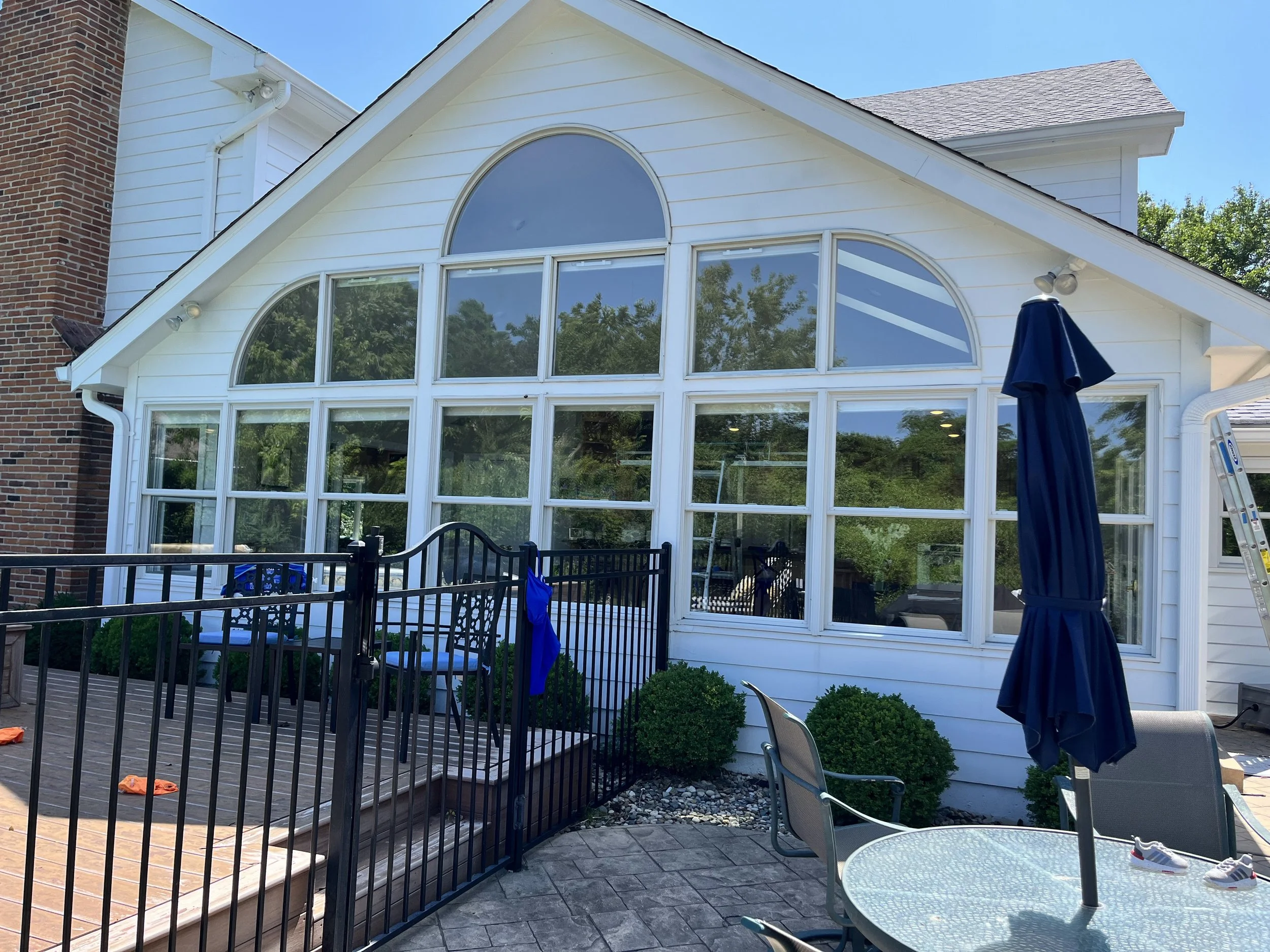 Backyard scene with a house featuring large arched windows, a wooden deck with a safety railing, patio furniture including a table and chairs, a closed patio umbrella, and a staircase leading up to the deck. The yard is enclosed by a black metal fenc