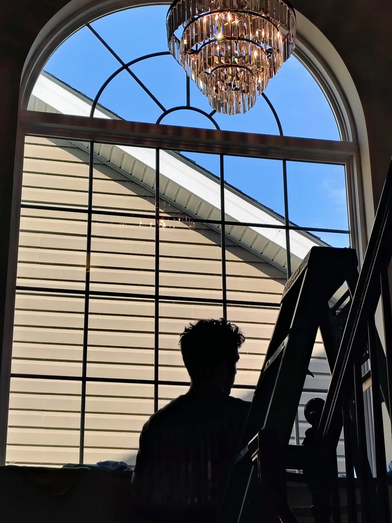Silhouette of a person sitting in front of a large arched window with a chandelier hanging above. Outside, there is a clear blue sky and part of the roof of a neighboring building visible.