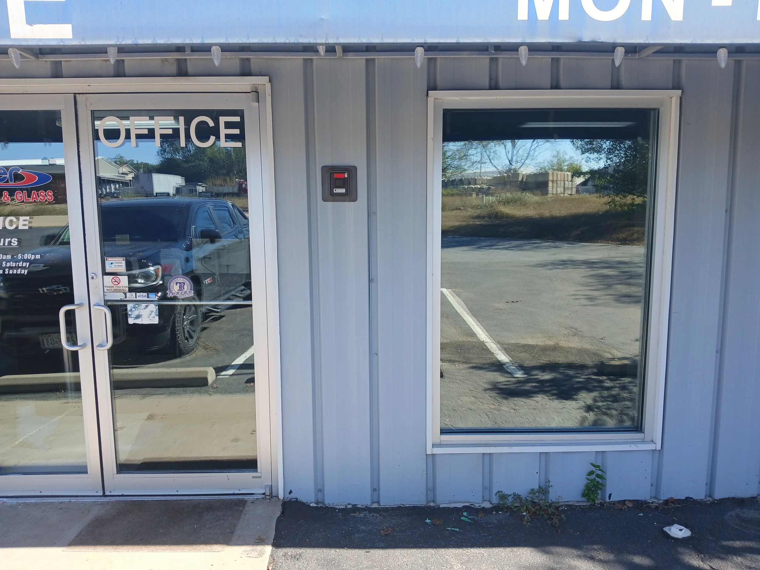 Empty storefront with glass double doors and large windows. 'OFFICE' sign on the door. Reflection of a black vehicle and buildings outside. Small plant growing near the base of the building.