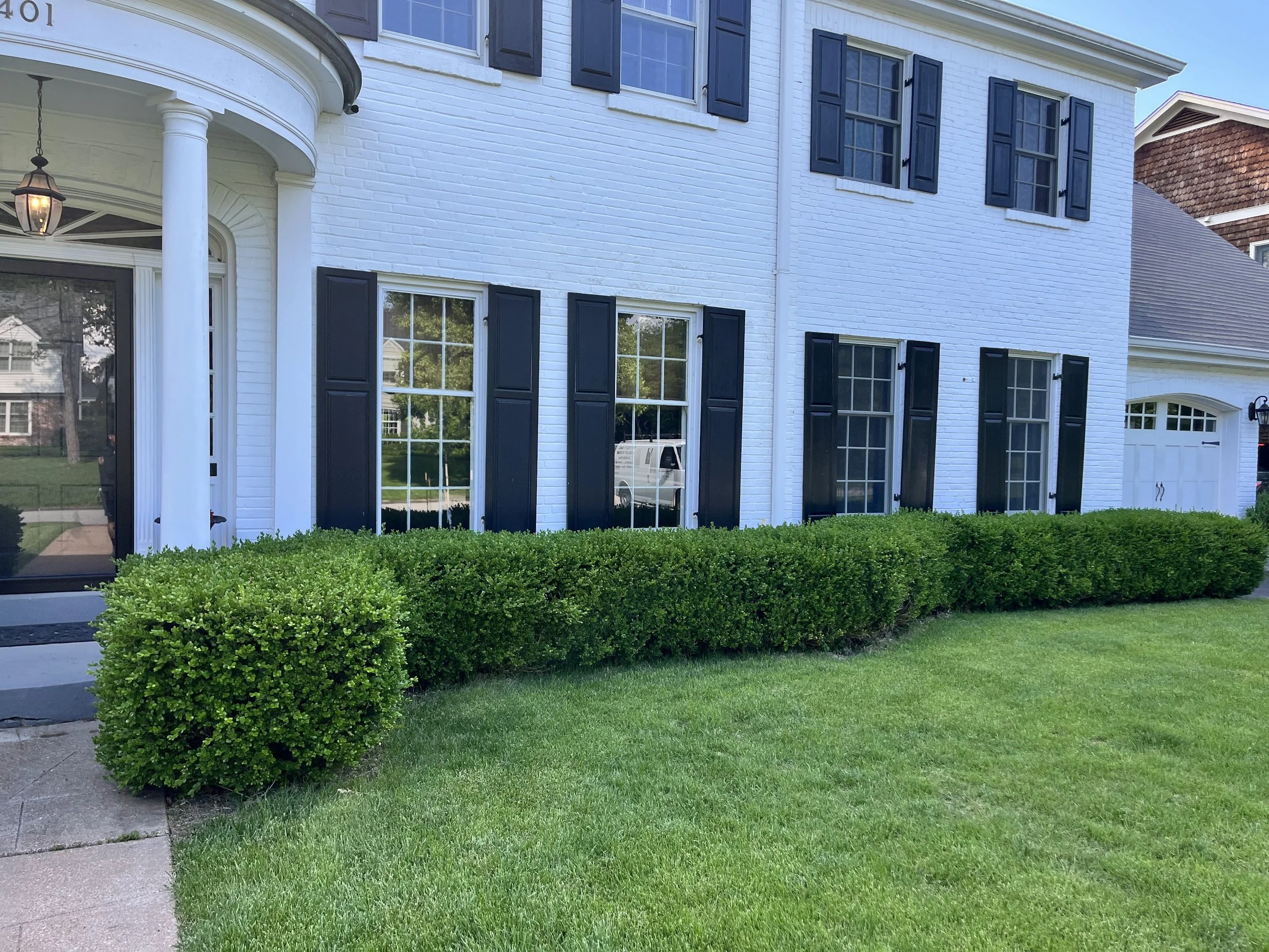 A white two-story house with dark blue shutters, a front porch with a hanging lantern, large windows, a garage, and a well-maintained green lawn with bushes.