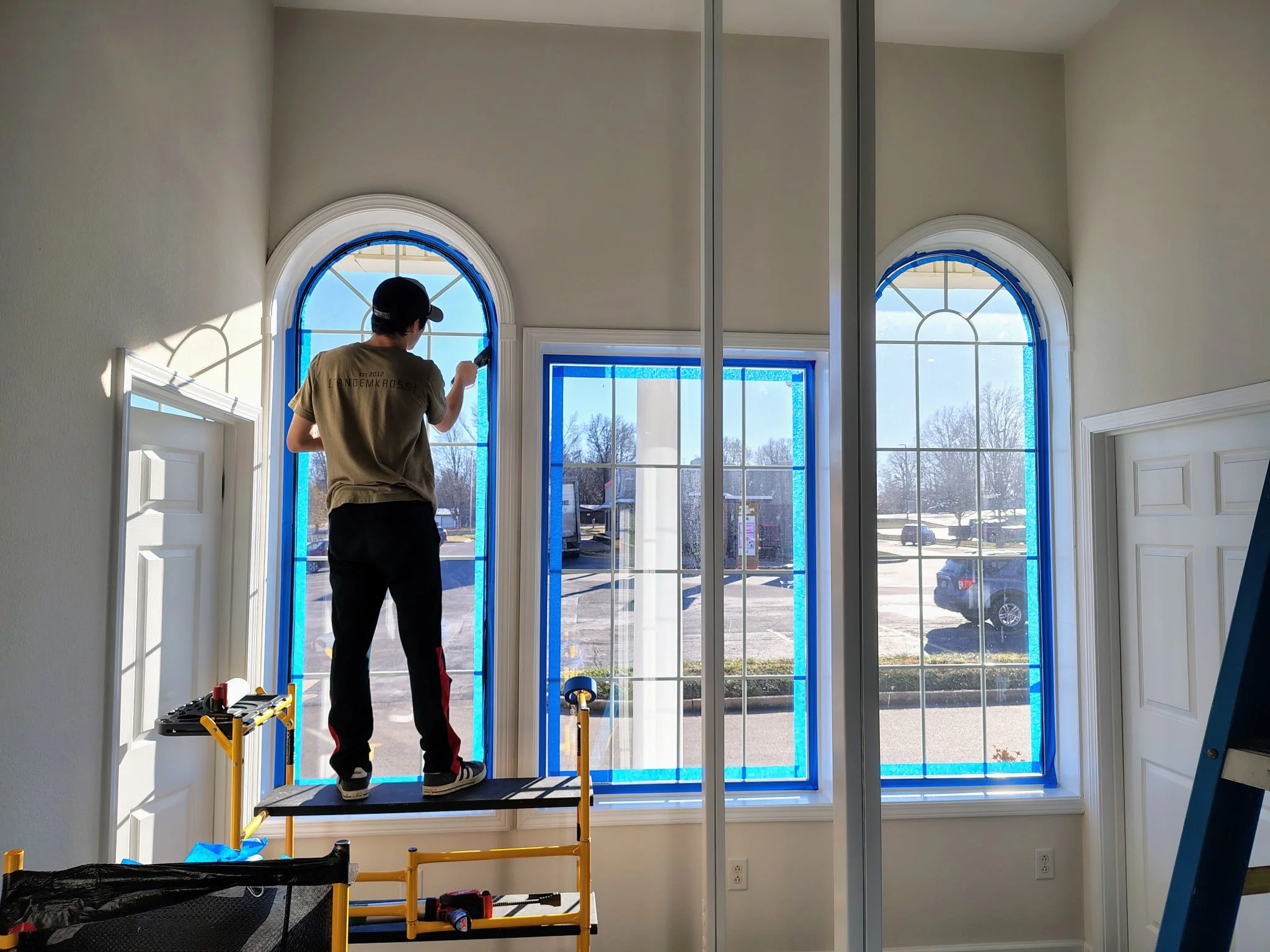 A person working on window installation or repair inside a house, standing on a small yellow scaffold. The house has large arched windows with blue tape around the frames, suggesting ongoing work. Outside, clear weather and parked cars are visible.