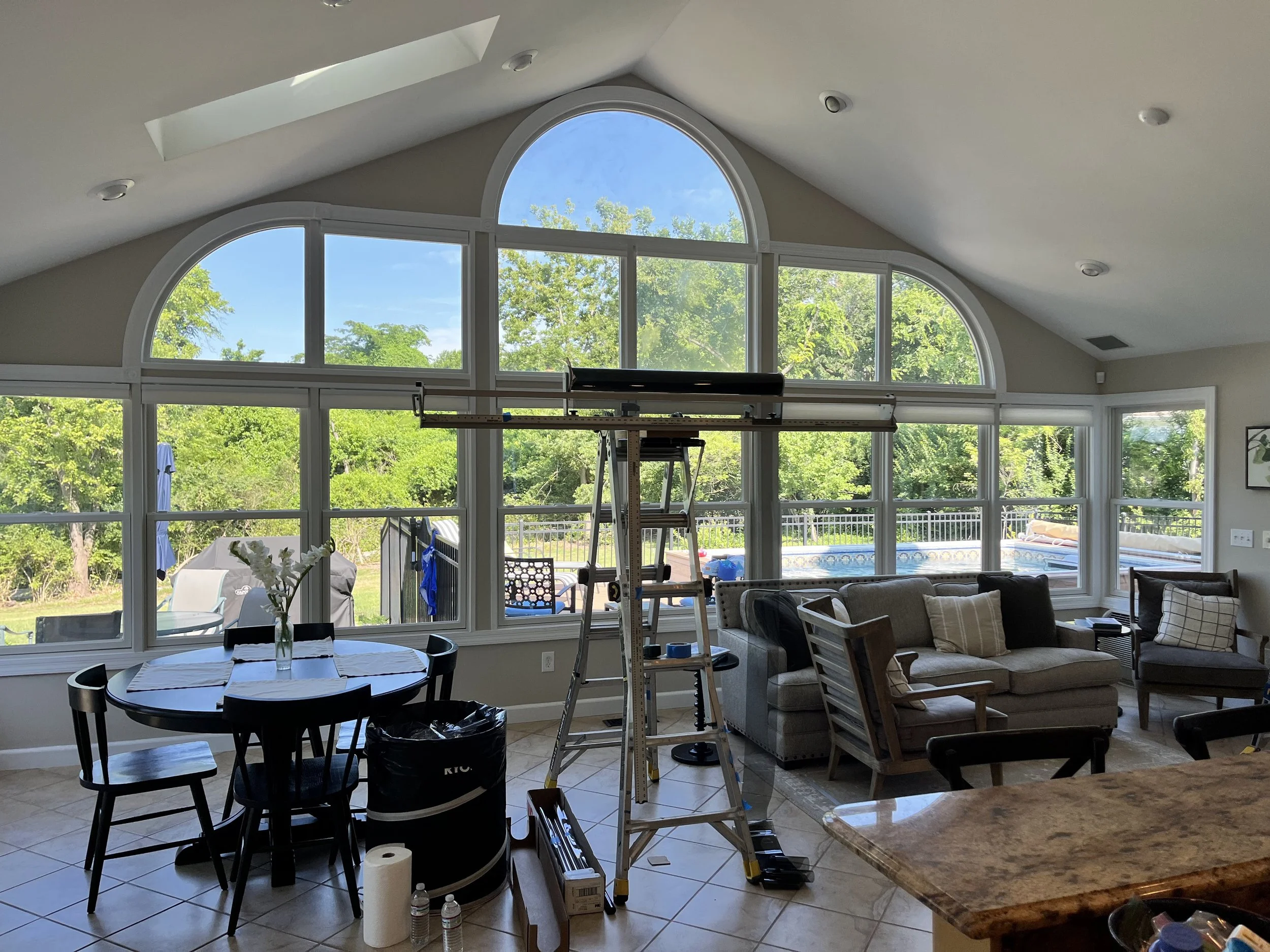 Living room with large arched windows showing a view of green trees and blue sky, furniture including a sofa, chairs, and a dining table, with various items and equipment such as a ladder, tools, and household supplies indicating ongoing renovations 
