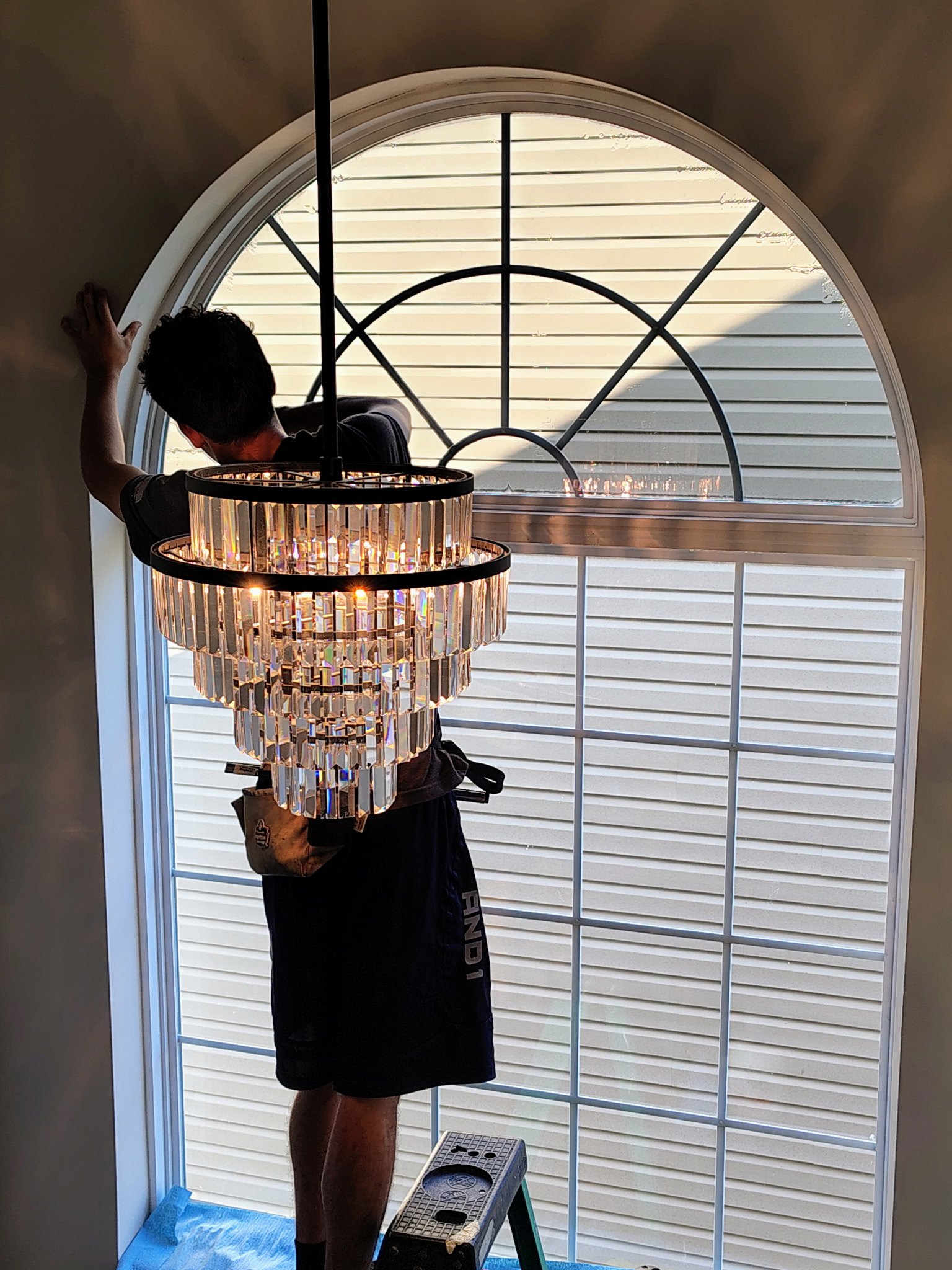 Person standing on a step stool, installing or adjusting a large arched window with horizontal blinds, during daytime.