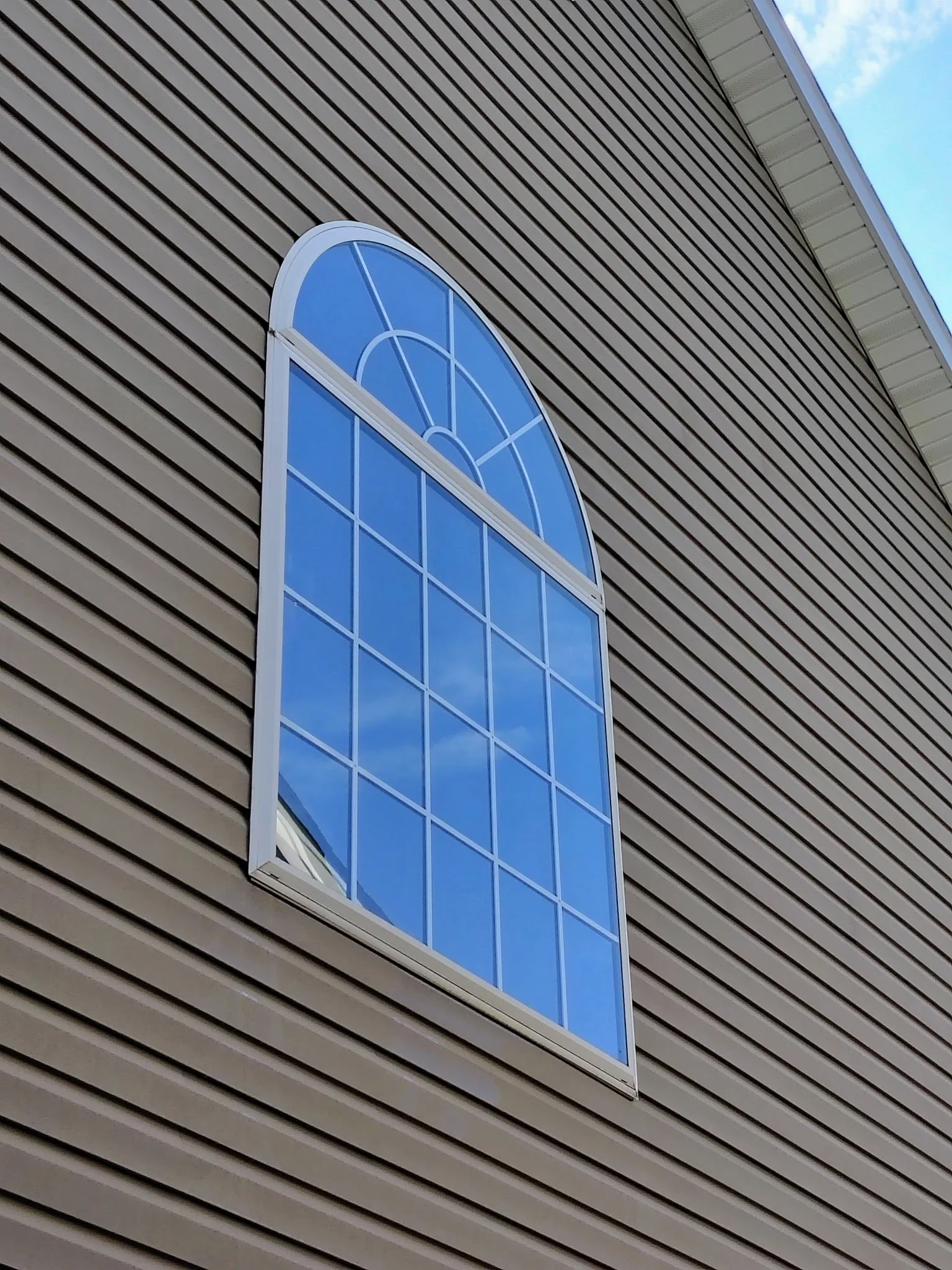 A large arched window with blue-tinted glass set into a building with horizontal beige siding. The sky with some clouds is reflected in the window.