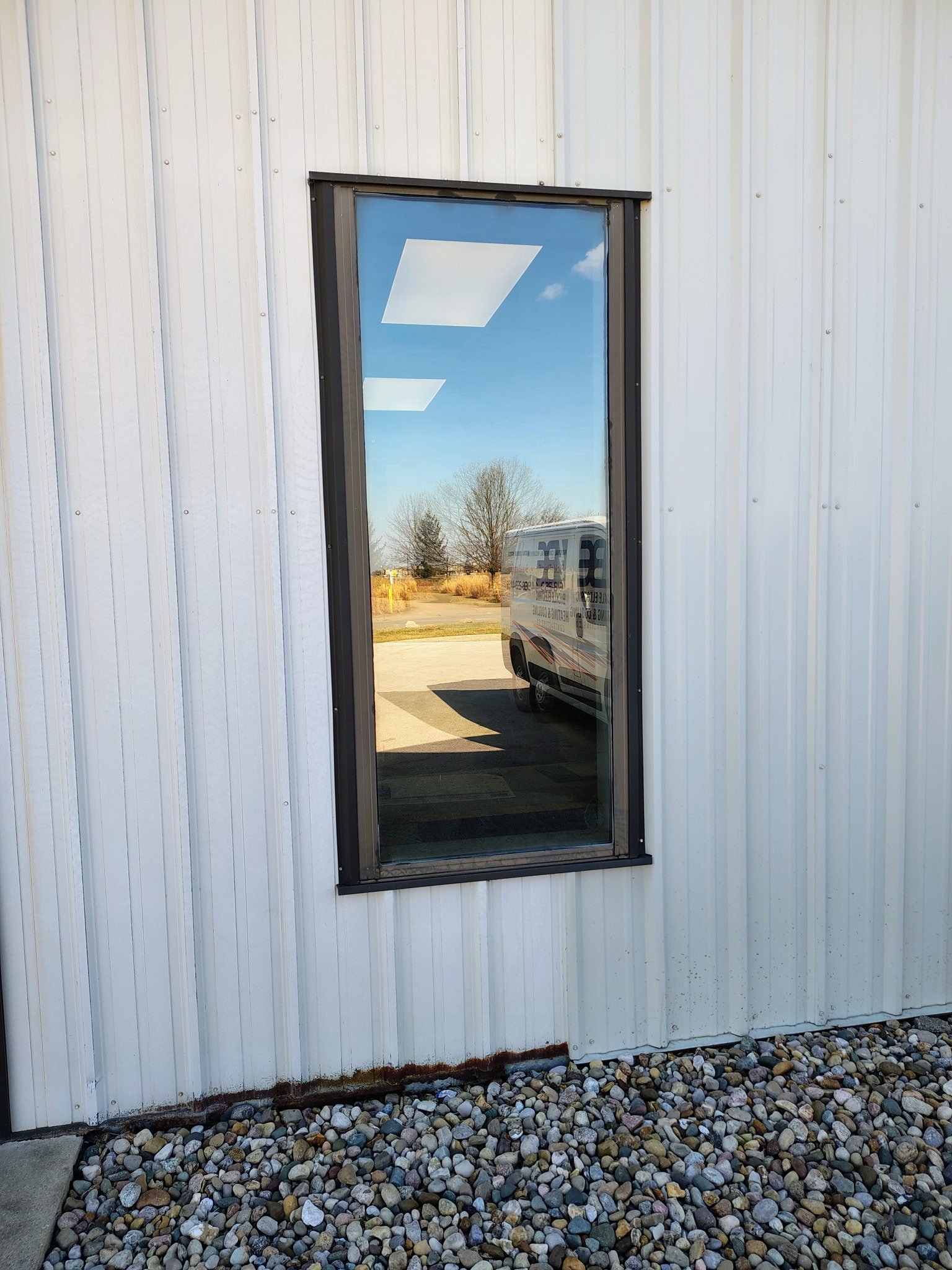 Exterior view of a building with white metal siding and a rectangular window with a black frame reflecting the blue sky, some clouds, a tree, and a van with writing on it, surrounded by a bed of small rocks.