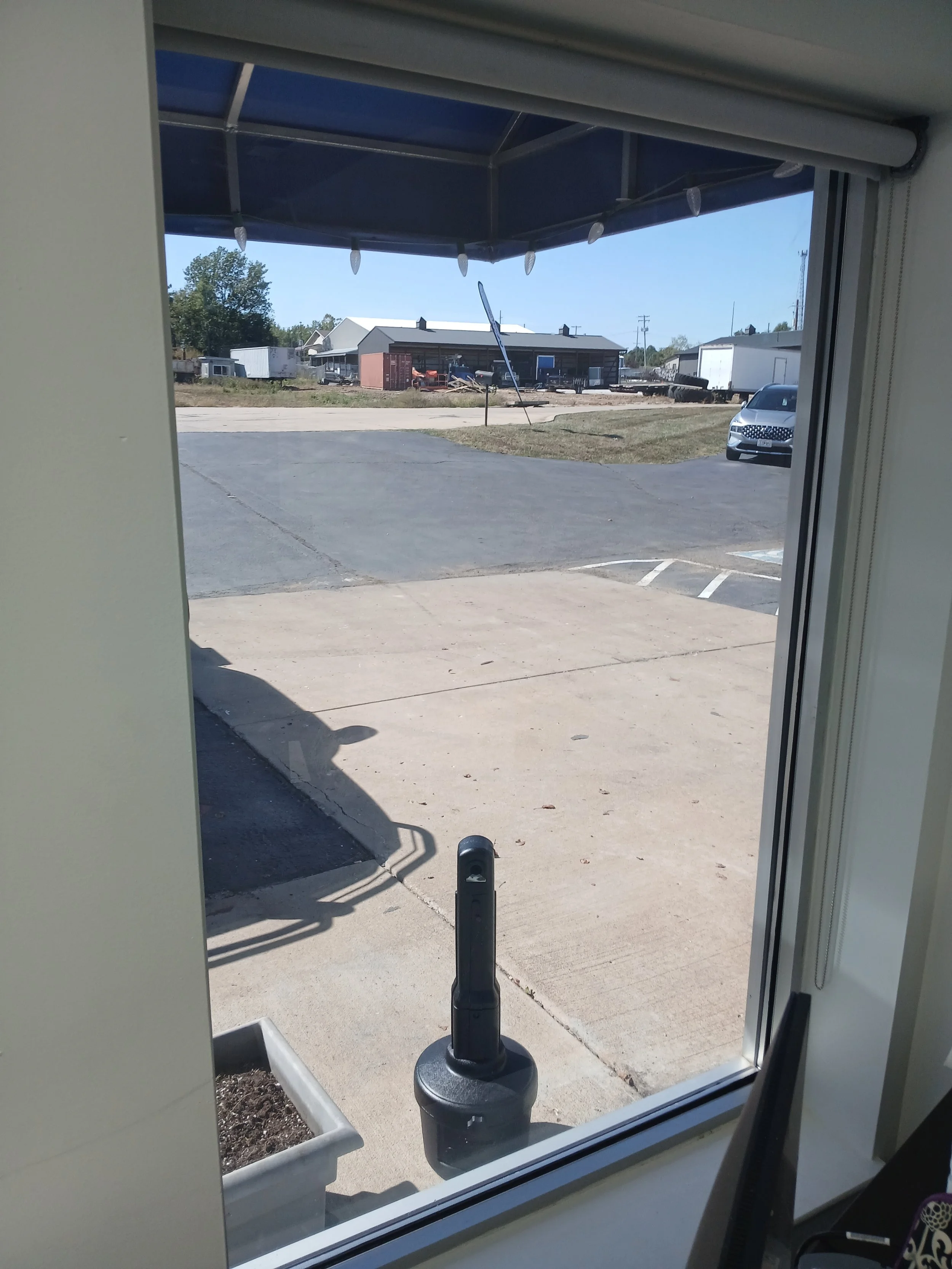 View of a parking lot with a gray car, a row of trailers and buildings, a clear blue sky, and a ladder leaning against a building, as seen through a window with a roller shade at the top.