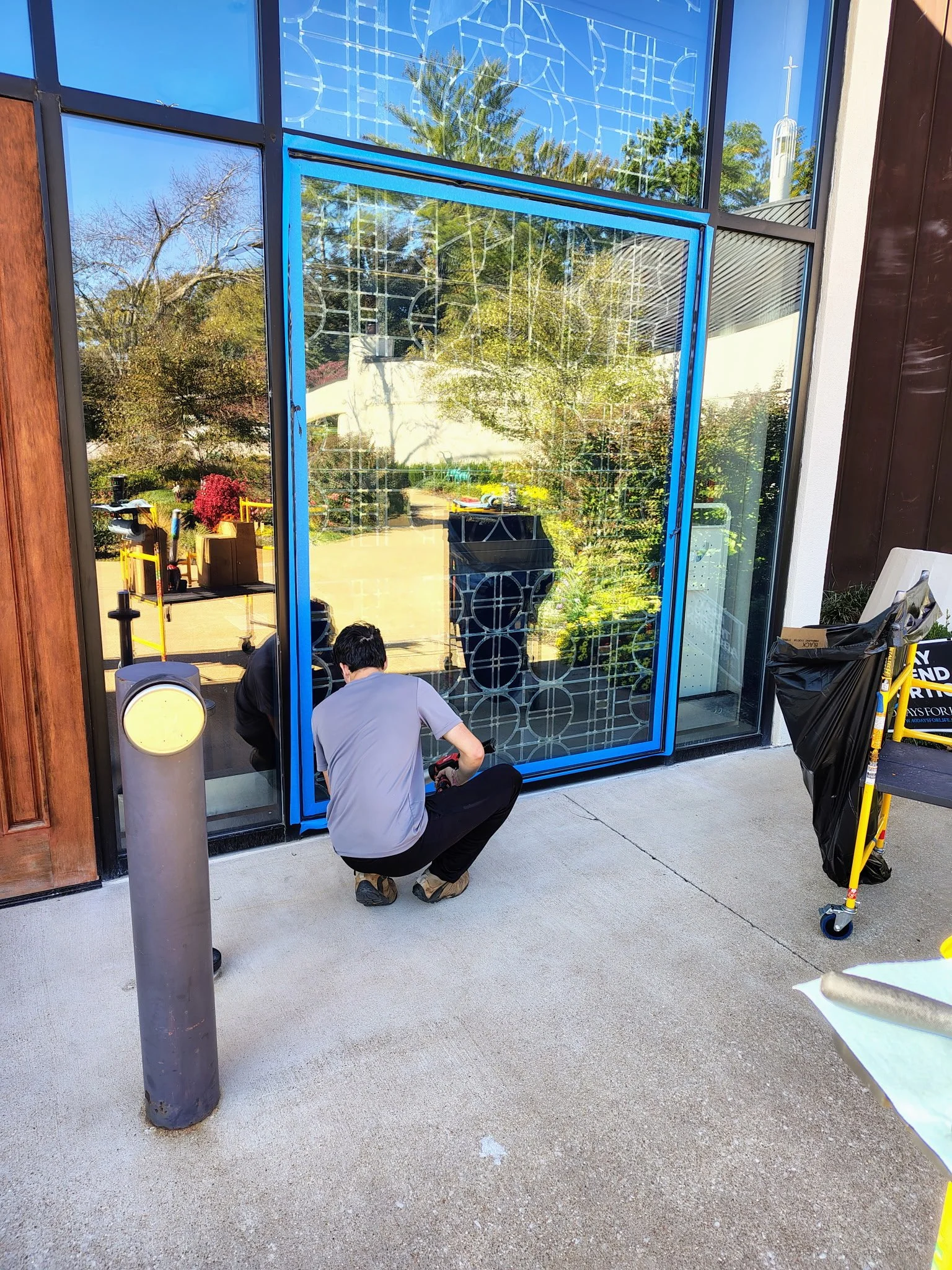 Two people working on the installation of a blue-framed glass door or window outside a building, with trees visible reflected in the glass.