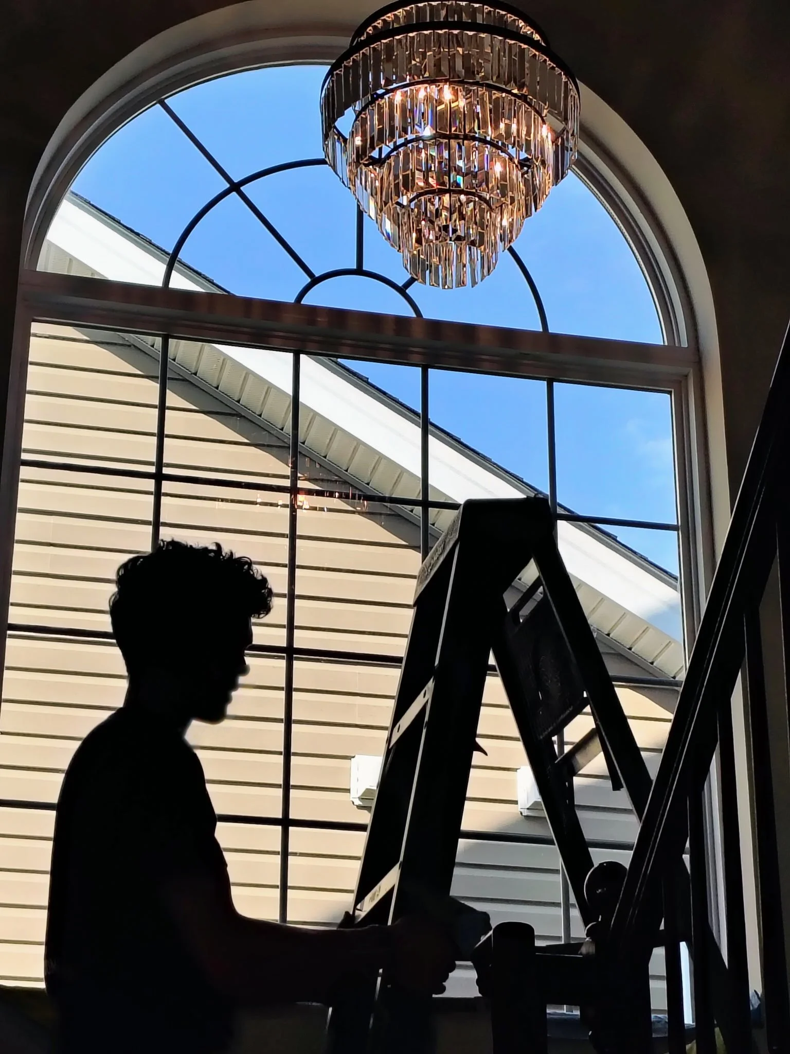 Silhouette of a person working on a laptop or computer inside building, with large arched window showing blue sky and part of house roof outside, chandelier hanging from ceiling.
