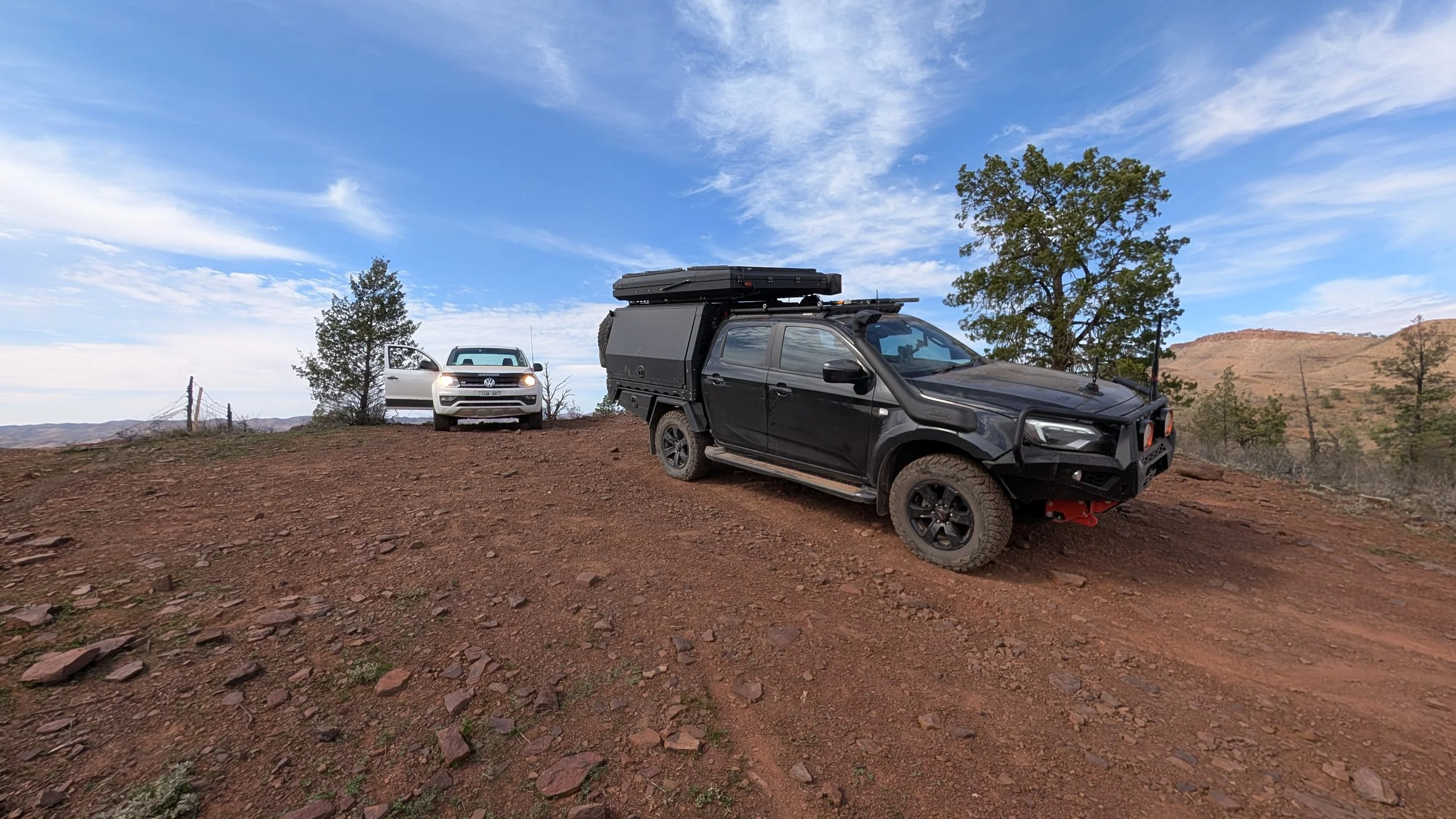 Two off-road vehicles parked on a dirt hill in a rugged landscape with trees and distant hills under a partly cloudy sky.