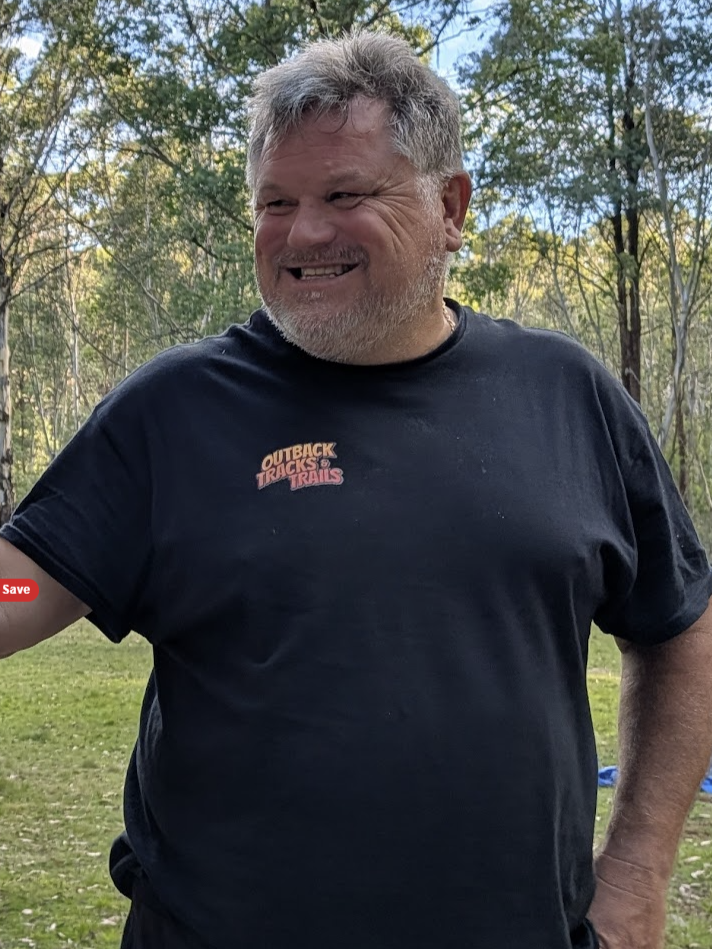 A smiling man with gray hair and beard outdoors in a wooded area, wearing a black t-shirt with a logo that says 'Outback Track & Trails'.
