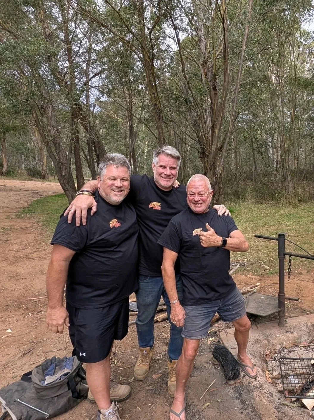 Three smiling men in black shirts with a logo are standing outdoors on a dirt trail with trees in the background. They are posing with arms around each other. One man points at the camera and is barefoot.