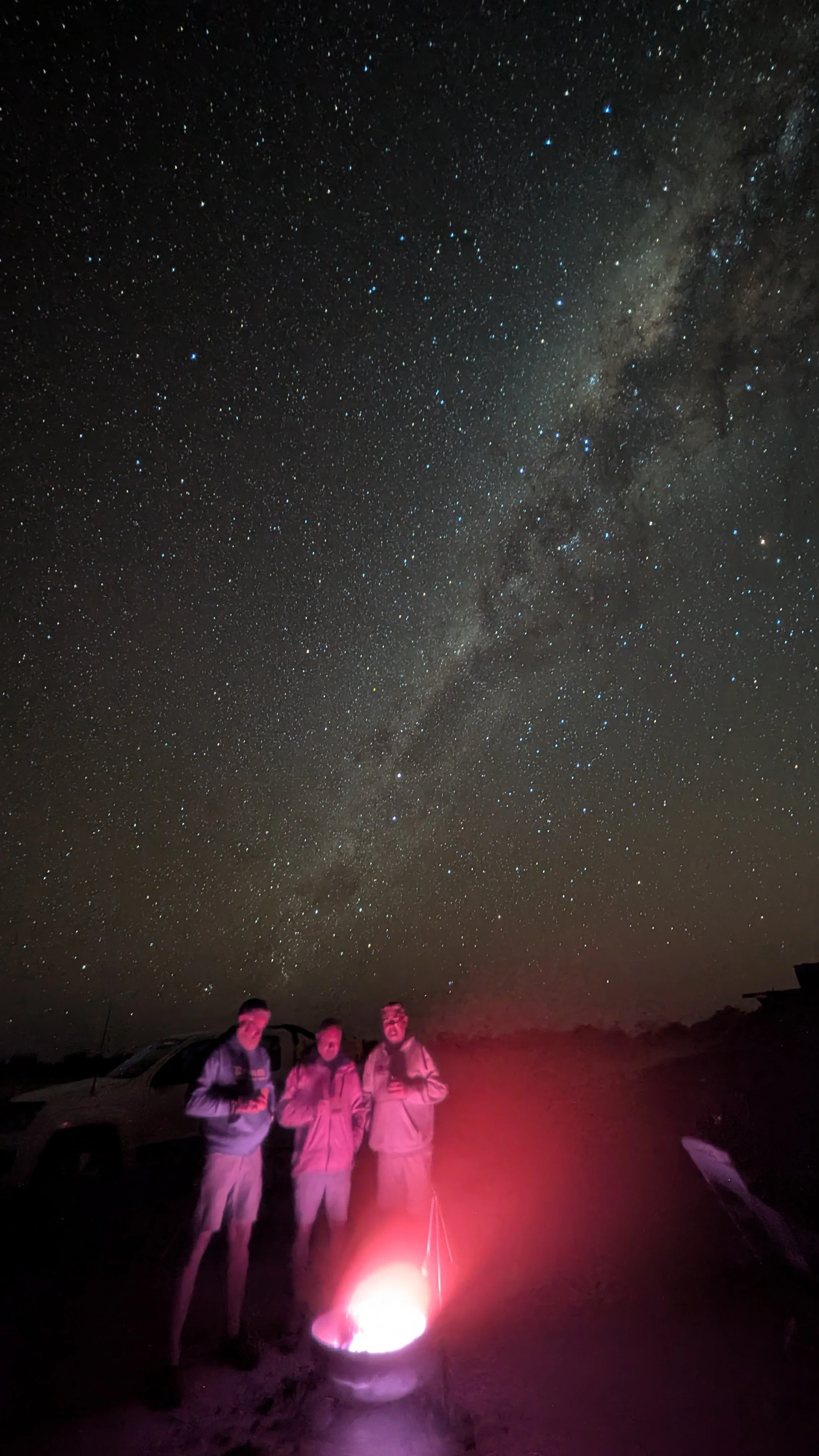 Three people standing outdoors at night around a bright campfire, with a clear, star-filled sky and the Milky Way galaxy visible above.