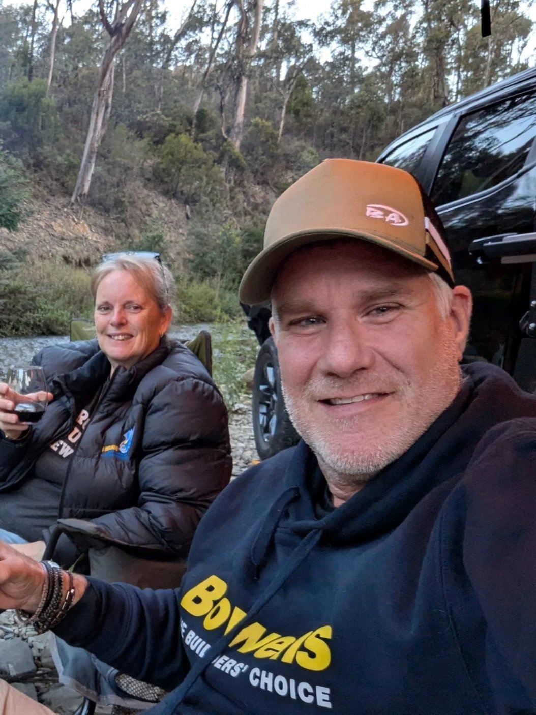 A man and a woman sitting outdoors near a river, smiling. The man is holding a glass of wine and wearing a cap and a dark hoodie, while the woman is wearing a black jacket and sunglasses. There are trees and a vehicle in the background.