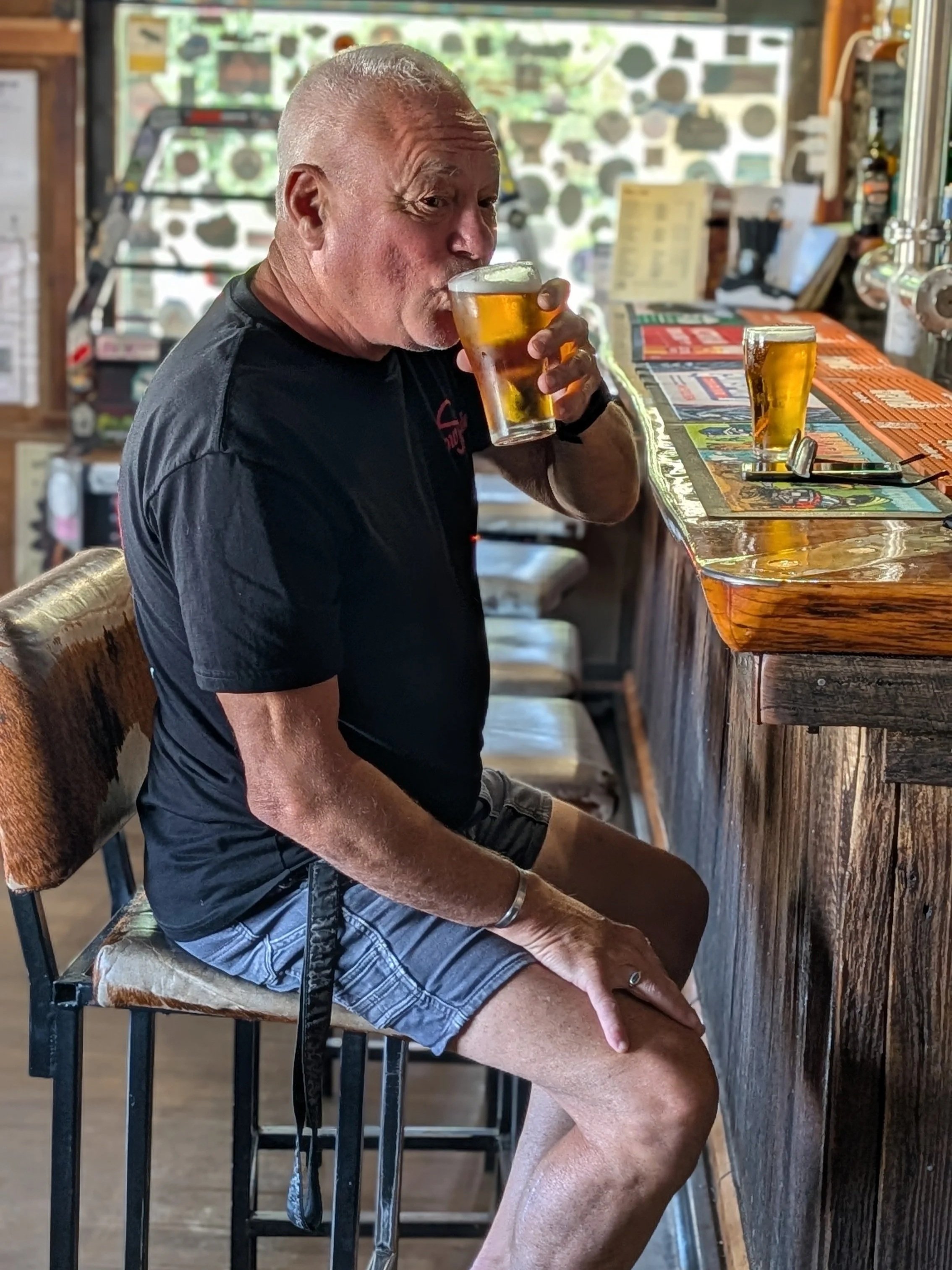 An older man with a shaved head sitting at a bar, drinking beer from a clear glass.