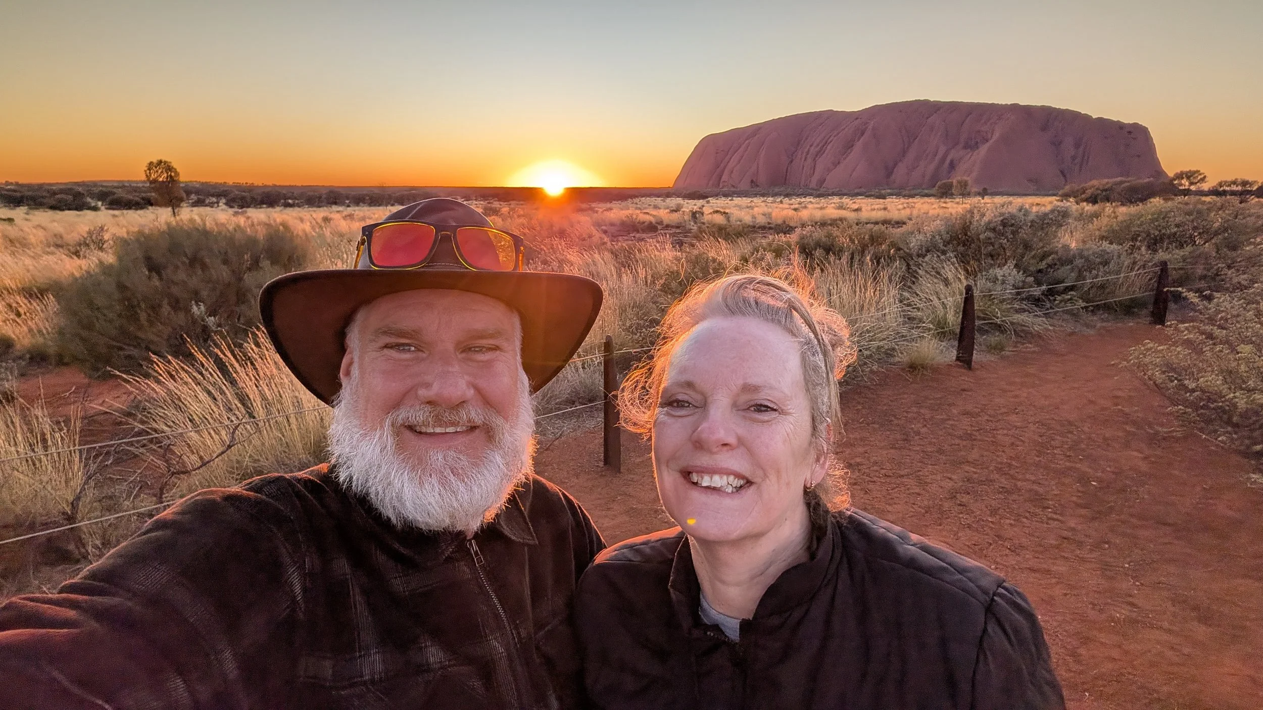 A smiling older man and woman taking a selfie at sunrise in a desert landscape with Uluru in the background, with dry grass and a dirt pathway.