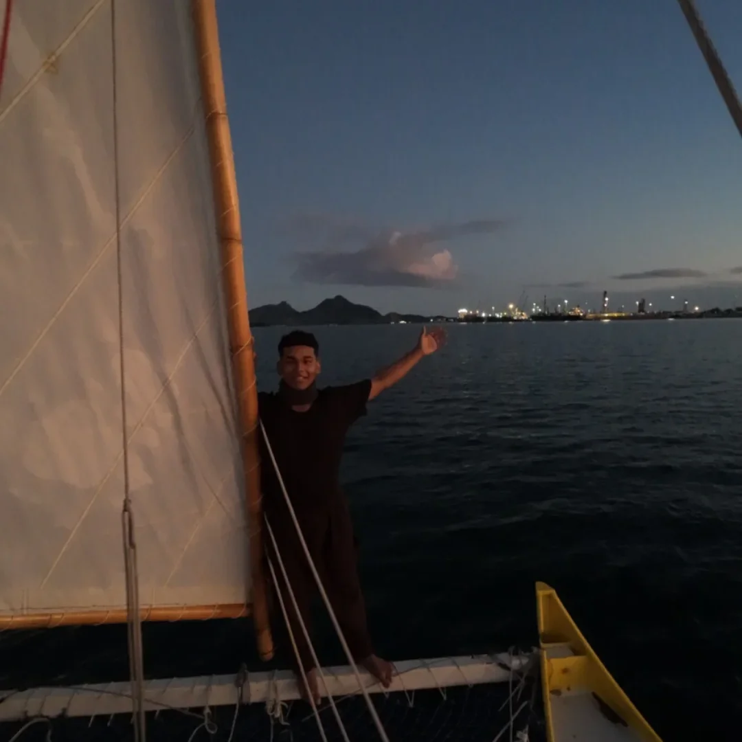 A young man standing on a boat at dusk, smiling and waving, with a view of the water, distant mountains, and city lights on the horizon.