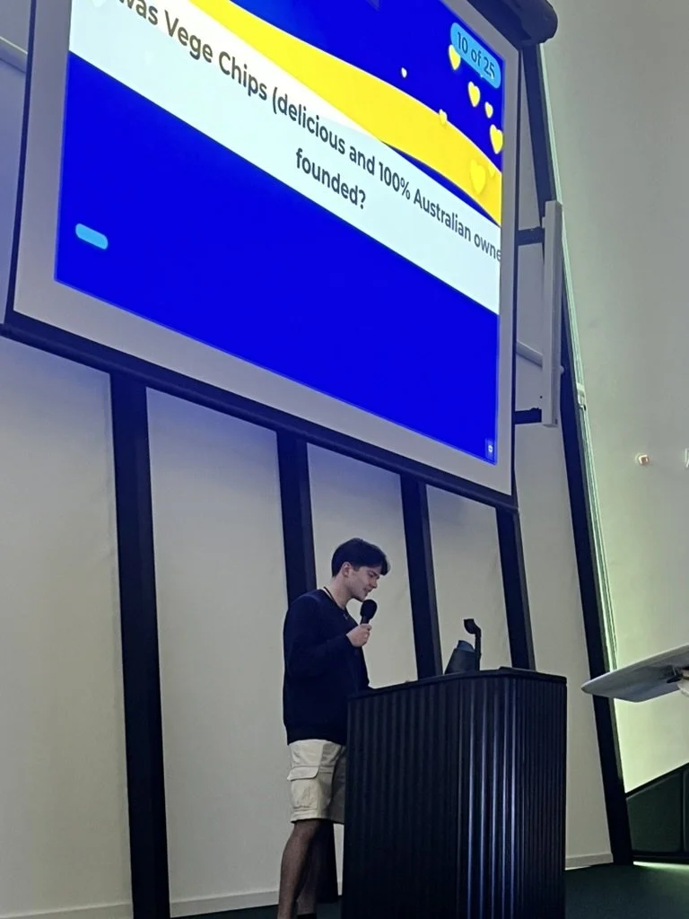 A young man standing at a podium with a microphone, giving a presentation in a room with large windows. A large screen behind him displays a question about Vegemite chips.