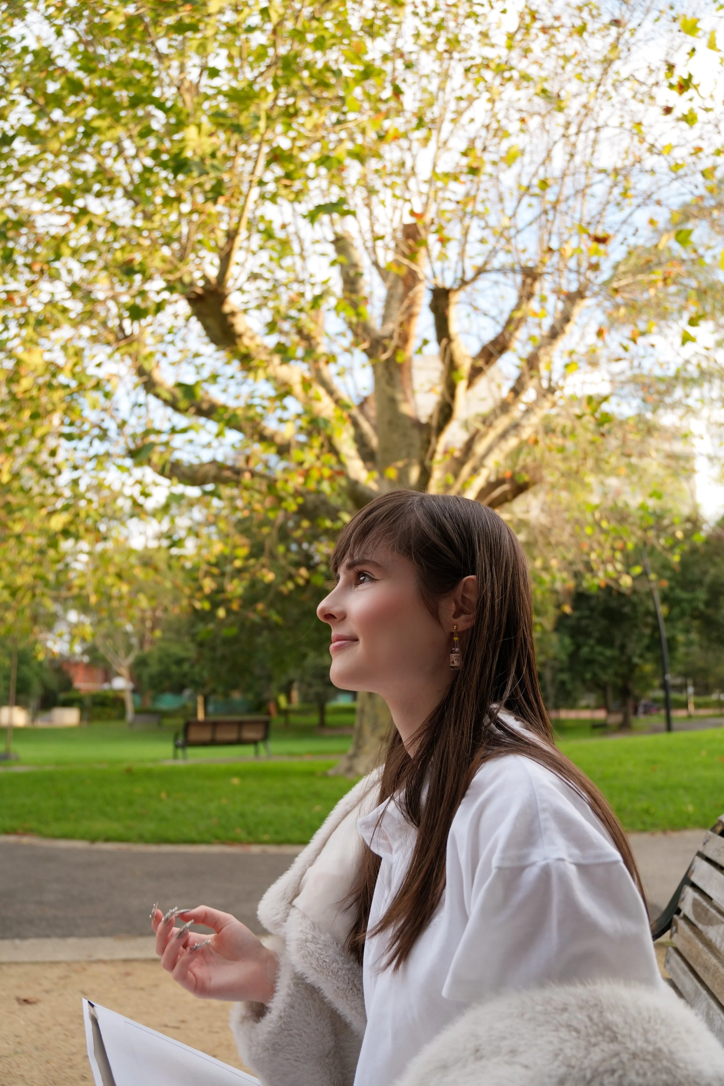 Side profile of a woman with long brown hair, wearing a white coat and earrings, sitting on a park bench in a park with trees and green grass, during the daytime.