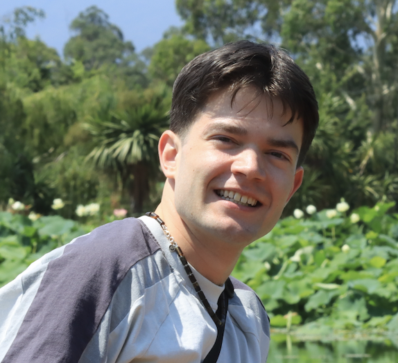 A young man (secretary) smiling outdoors near a pond with lily pads and greenery in the background.