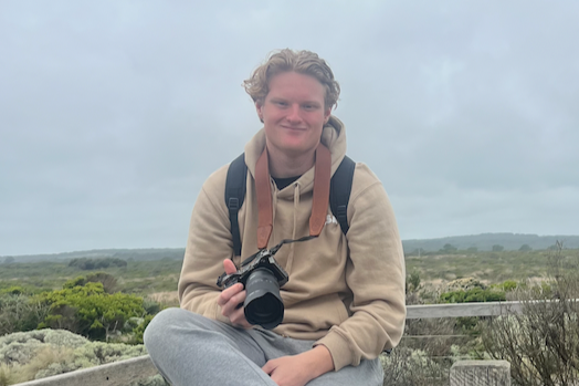 A young man (Events Manager)  with curly hair sitting outdoors on a cloudy day with a backpack, holding a camera, and smiling at the camera.