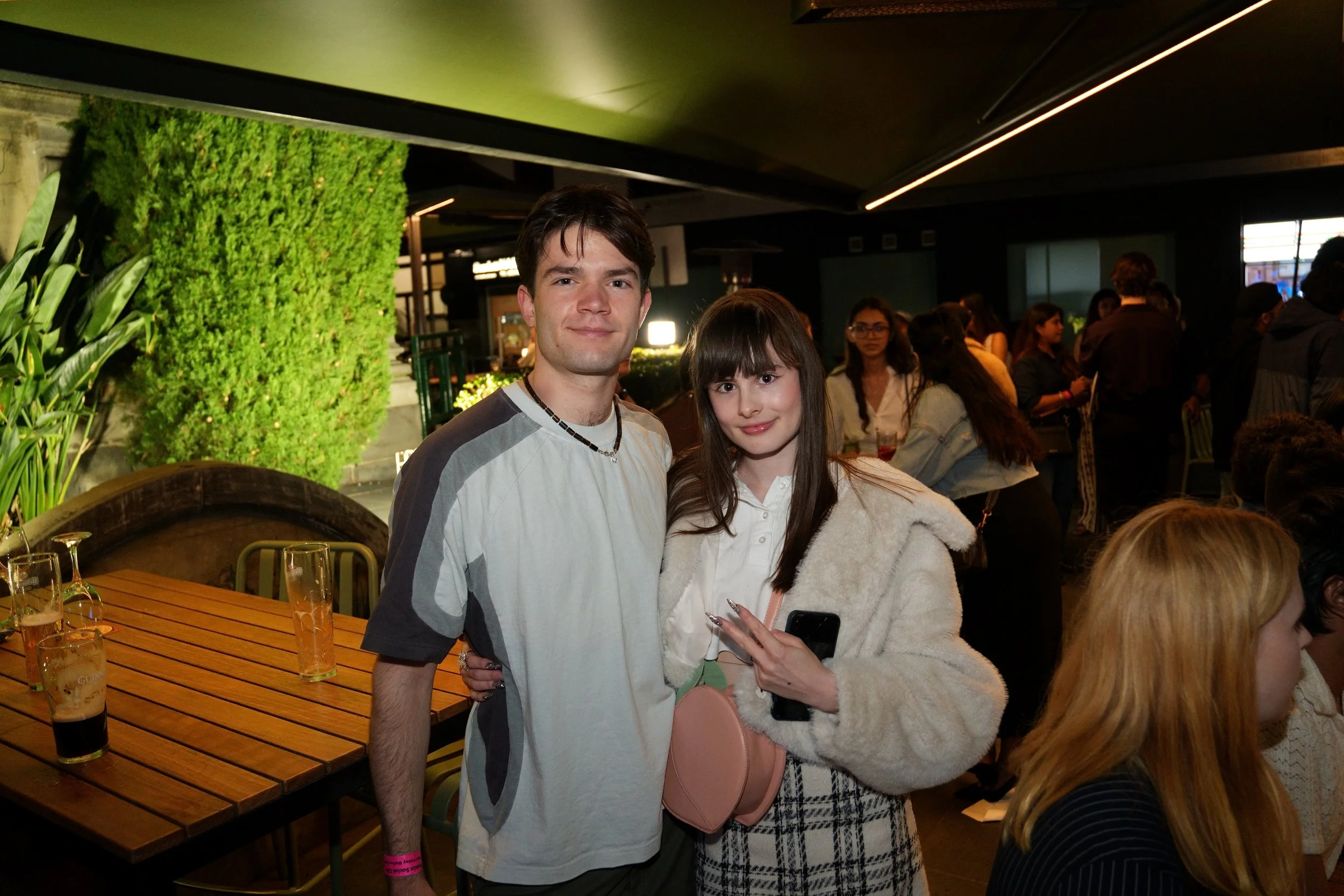 Two people, a young man and young woman, are posing together at a social gathering indoors. The man has short dark hair, is wearing a light gray and white shirt with a black necklace, and has a pink wristband. They are standing close with the woman's arm around the man's waist. The woman has long dark hair with bangs, is wearing a beige fuzzy jacket, and is holding a black phone and a pink purse. There are several people in the background and glasses of beer on the table.